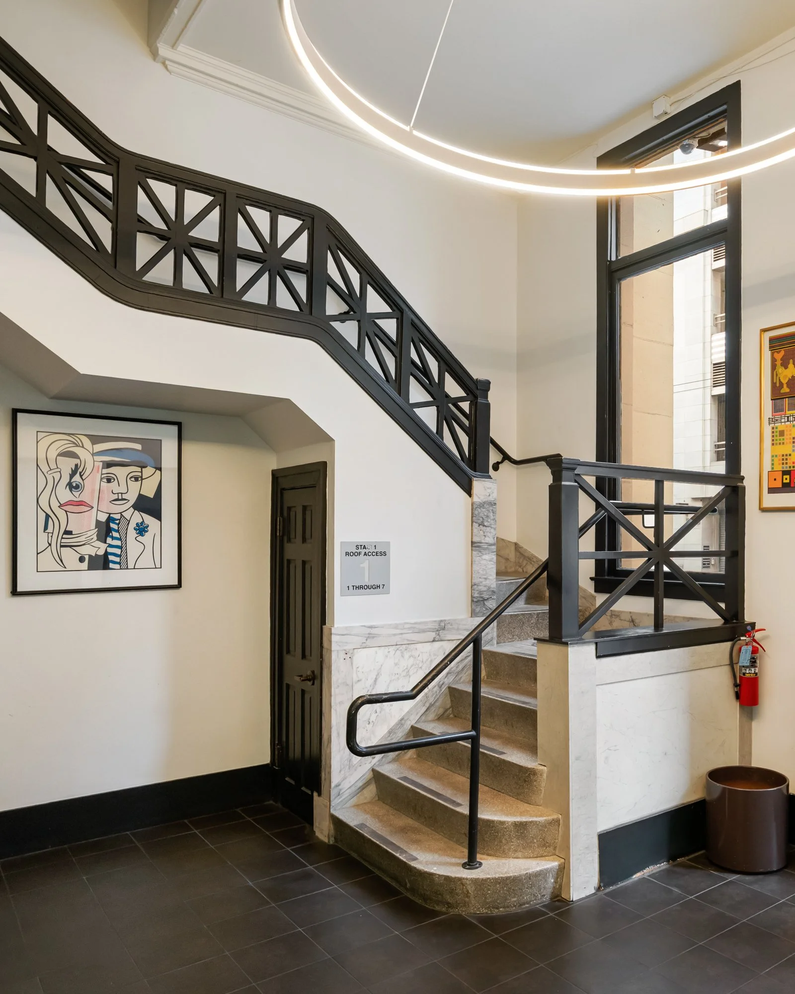 Interior of a building with stairs, black railings, and large windows. Artwork on the wall and a fire extinguisher near the stairs.