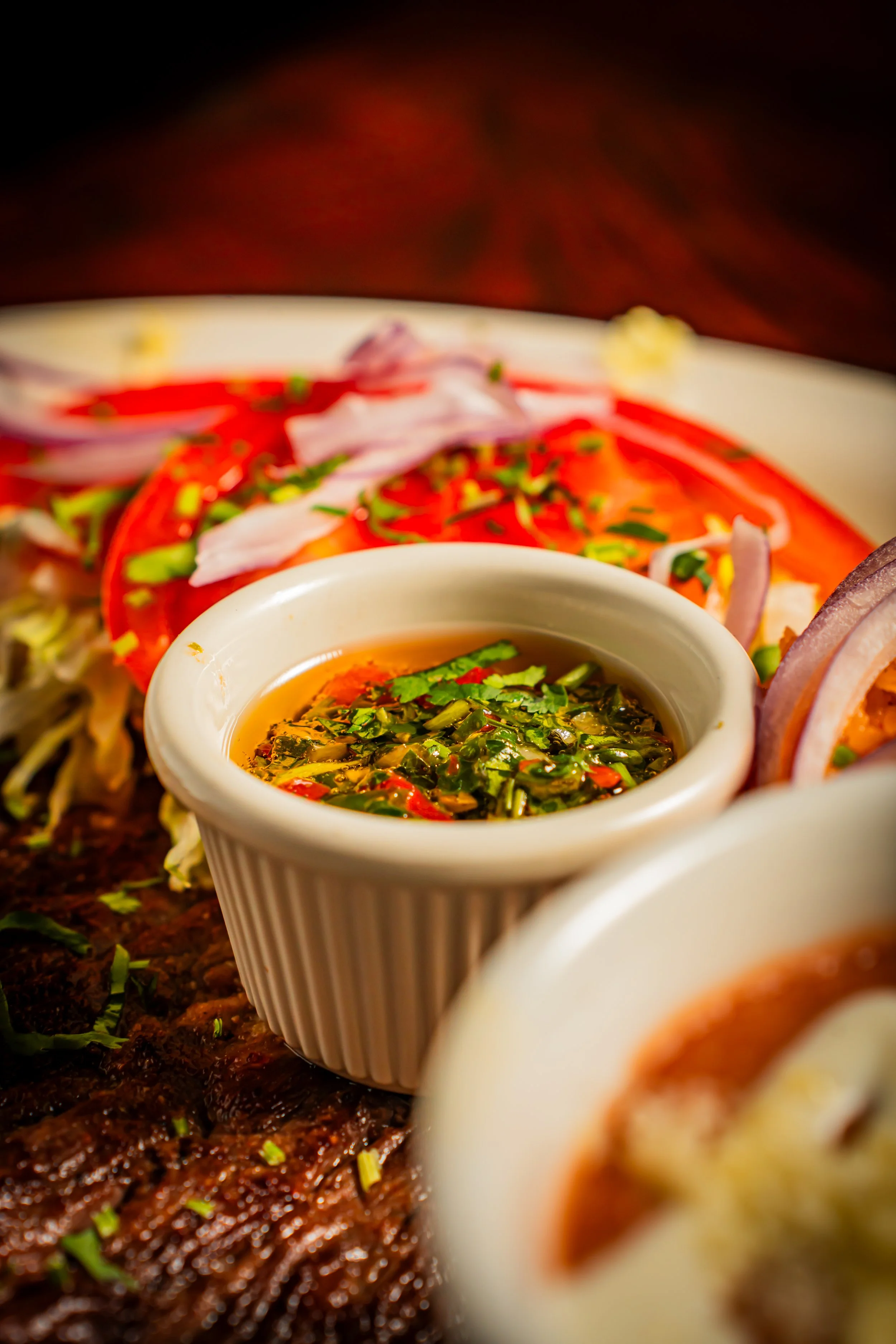 Close-up of Mexican food with colorful toppings, a small cup of sauce, and grilled meat on a wooden surface.
