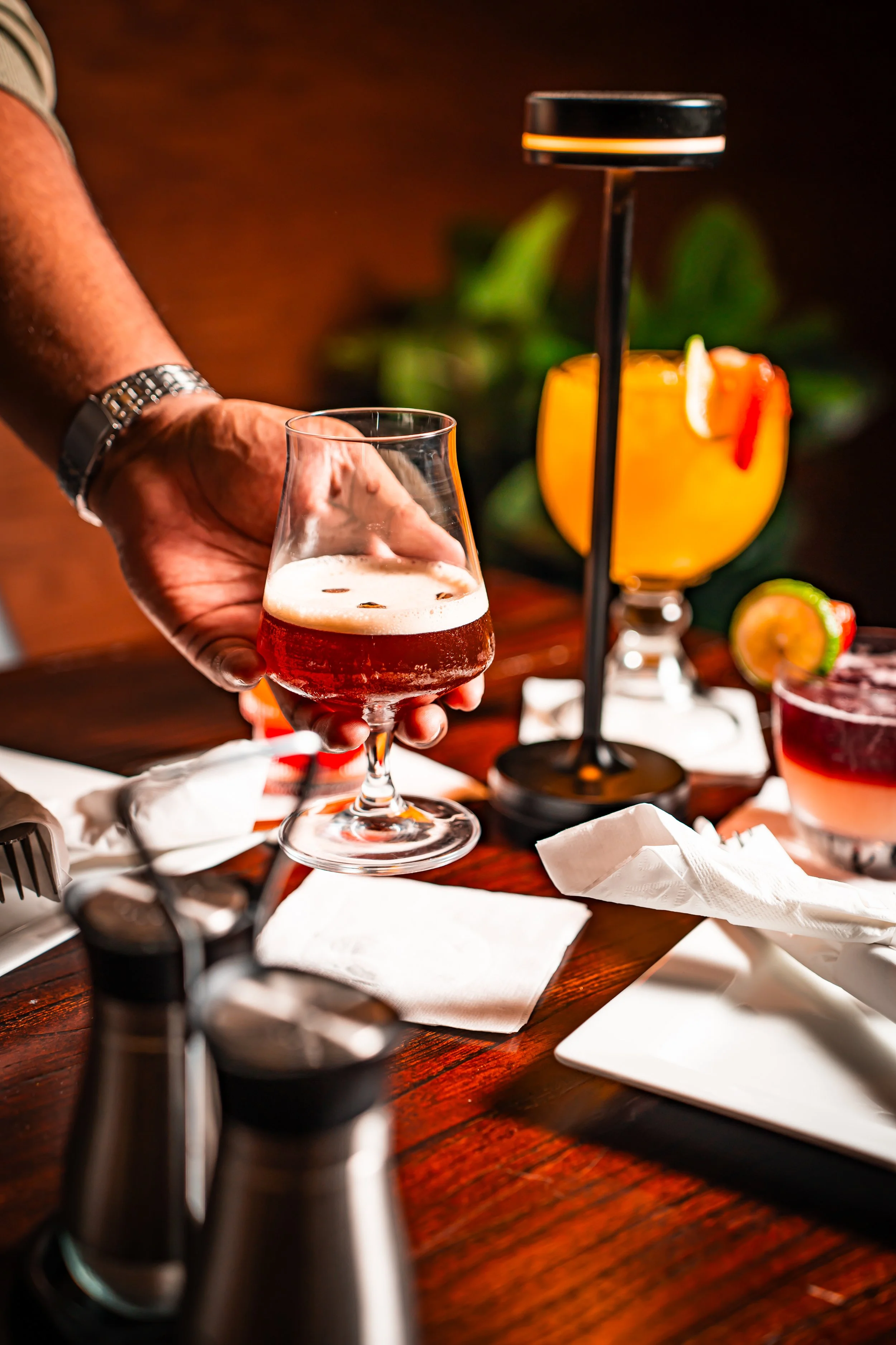 Person with a wristwatch holding a glass of reddish-amber colored beer with a foamy head, set on a wooden table with napkins, salt and pepper shakers, and various cocktails in the background.