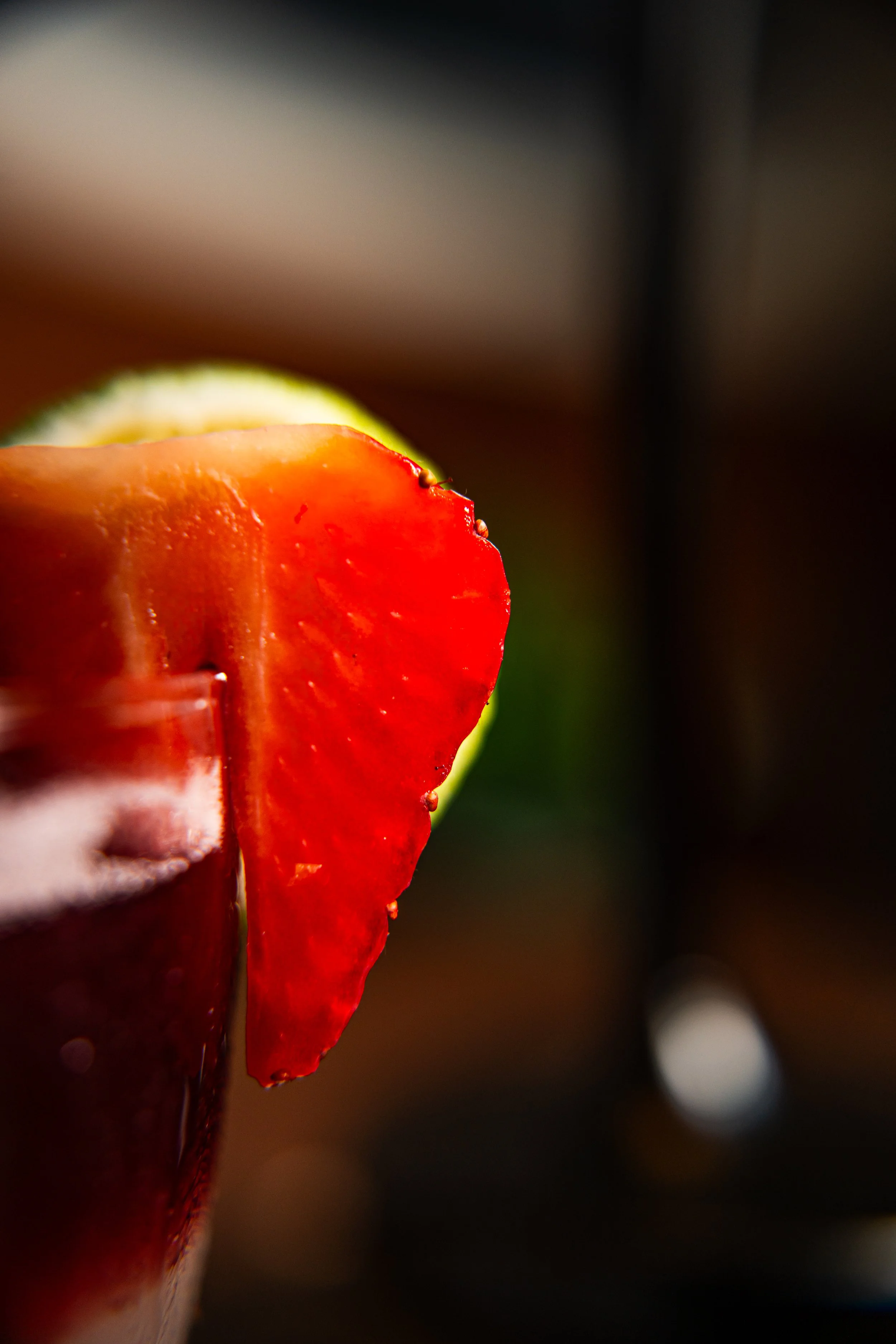 Close-up of a red apple slice on the rim of a dark glass drink, with a blurred background.