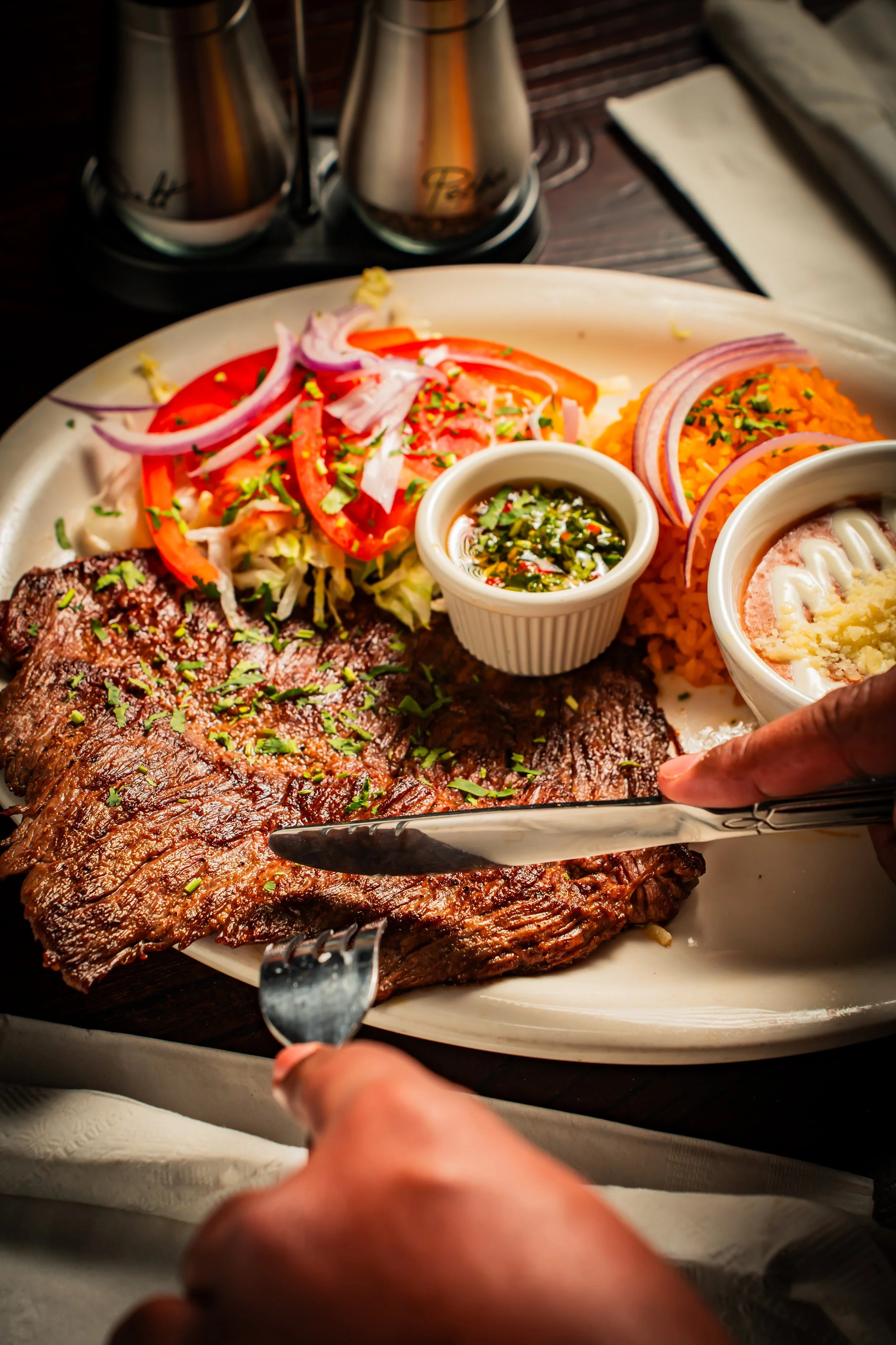 A plate of grilled steak with chopped herbs, side salad with tomato, onion, and lettuce, two bowls of rice and beans, and a small dish of sauce, on a dark wooden table.