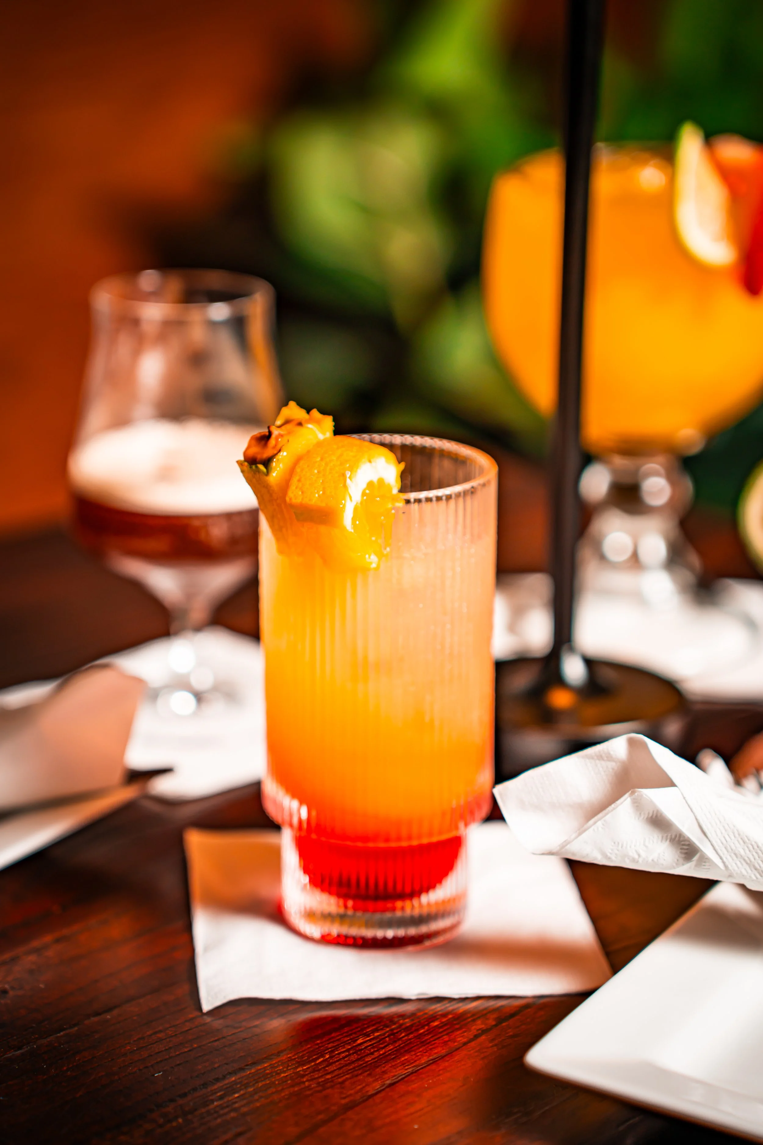 A colorful cocktail garnished with orange peel and a cherry, served on a white napkin on a wooden table.