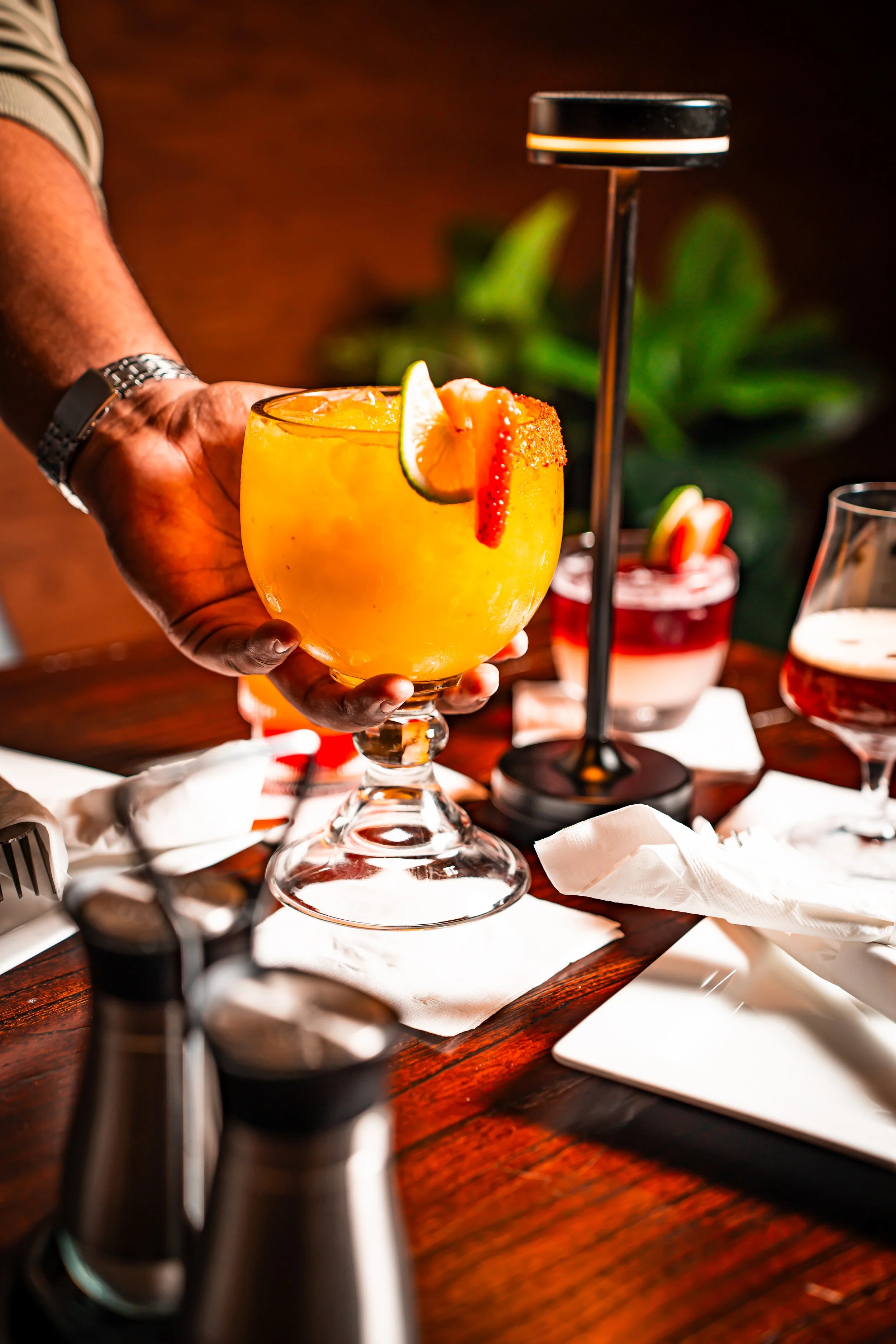 A person holding a large glass of orange cocktail garnished with a lime wedge and a chili pepper on an elegant wooden table with other drinks and tableware.