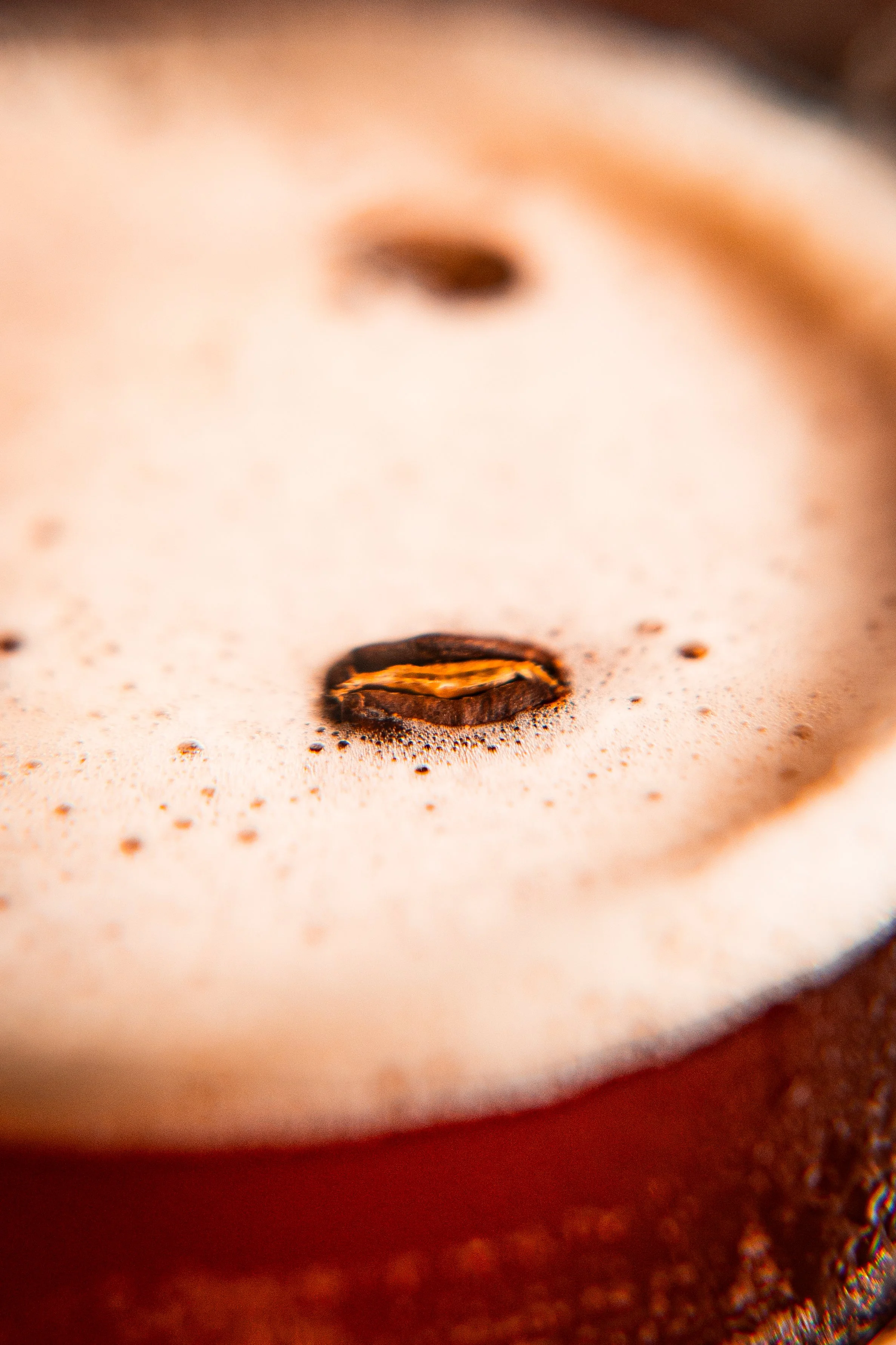Close-up macro shot of coffee bean on a surface resembling a face, with foam or froth creating cheeks and lips, and a blurred background.