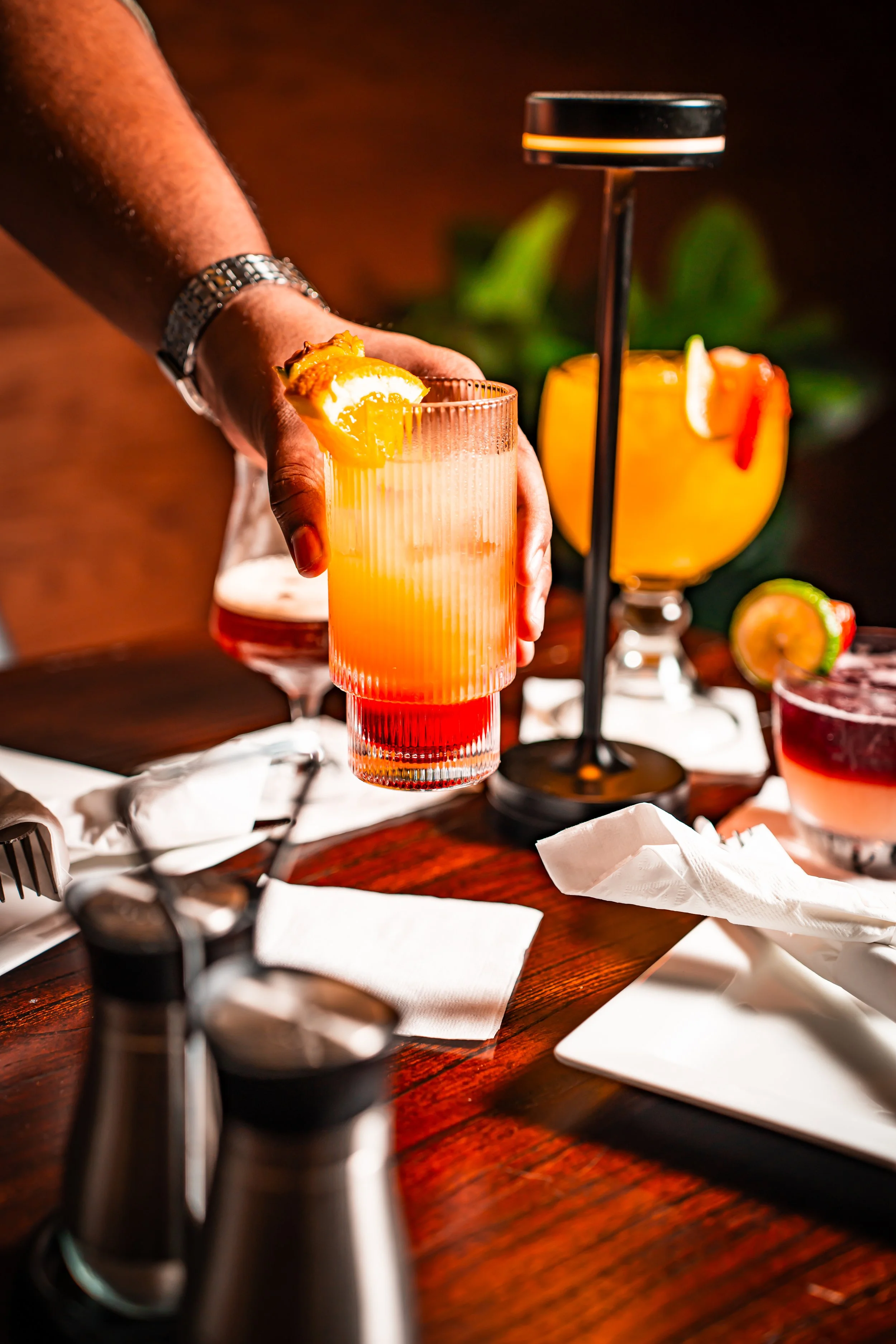 Close-up of a bartender's hand holding a colorful cocktail garnished with an orange slice. Several other drinks and bar tools are visible on the wooden bar counter.