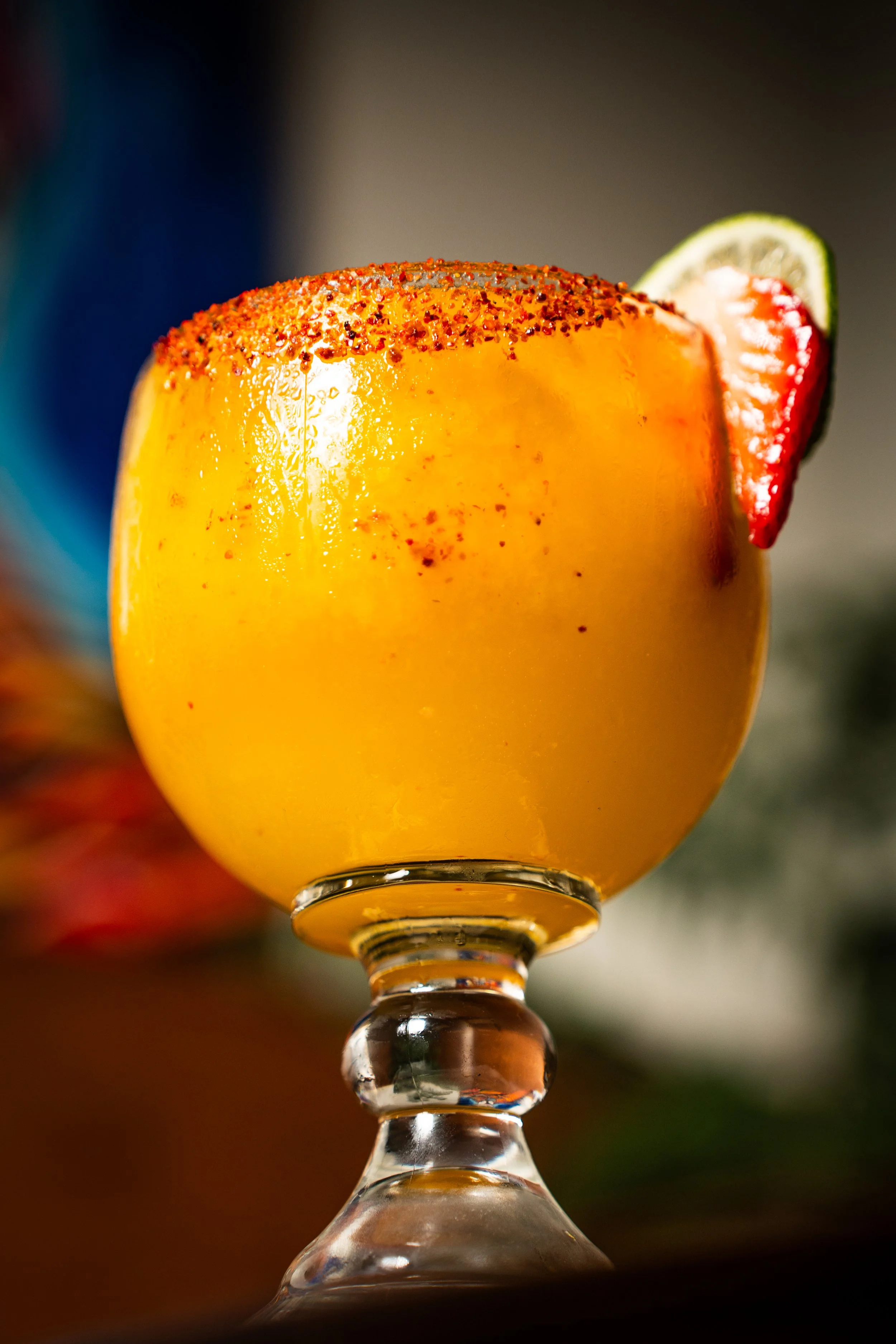 A close-up of a yellow mango smoothie in a glass, garnished with a lime and strawberry slice on the rim, with a blurred background.