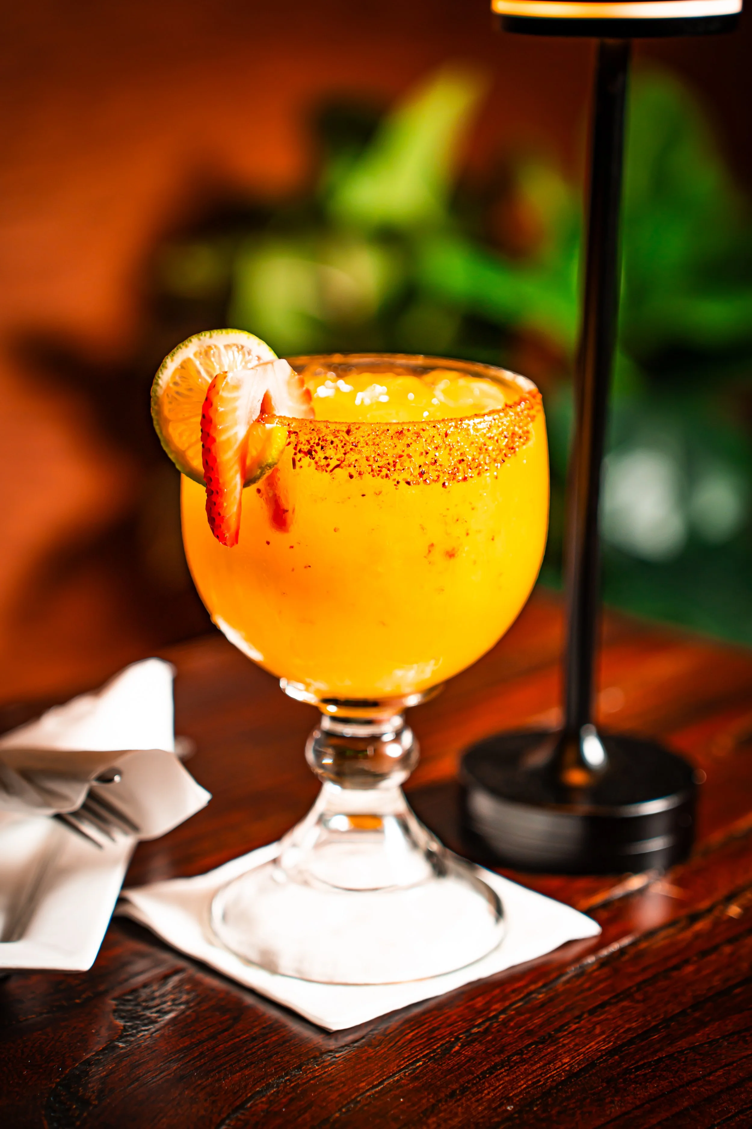 A vibrant yellow cocktail garnished with lemon and strawberry slices, served in a glass with a salted rim on a napkin, with a blurred background and a lamp.
