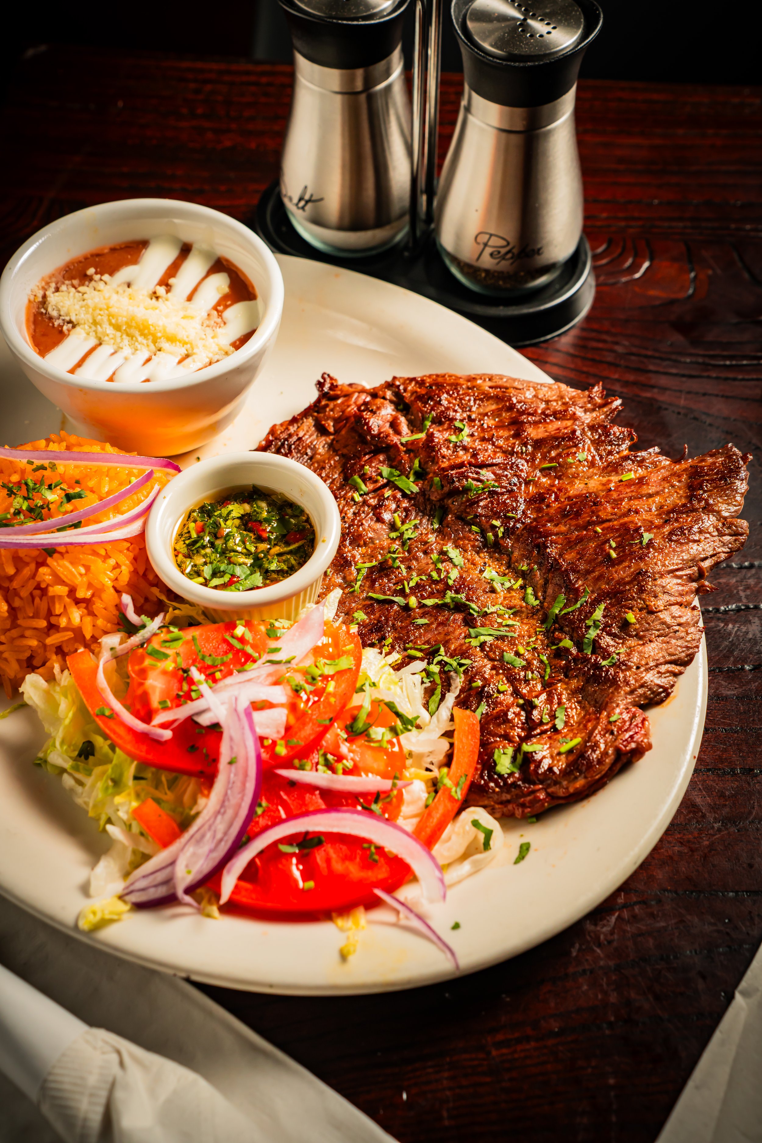 Plate of grilled steak with Mexican rice, fresh salad with tomato, onion, and cilantro, and sides including salsa and sour cream in a bowl, with salt and pepper shakers in the background.
