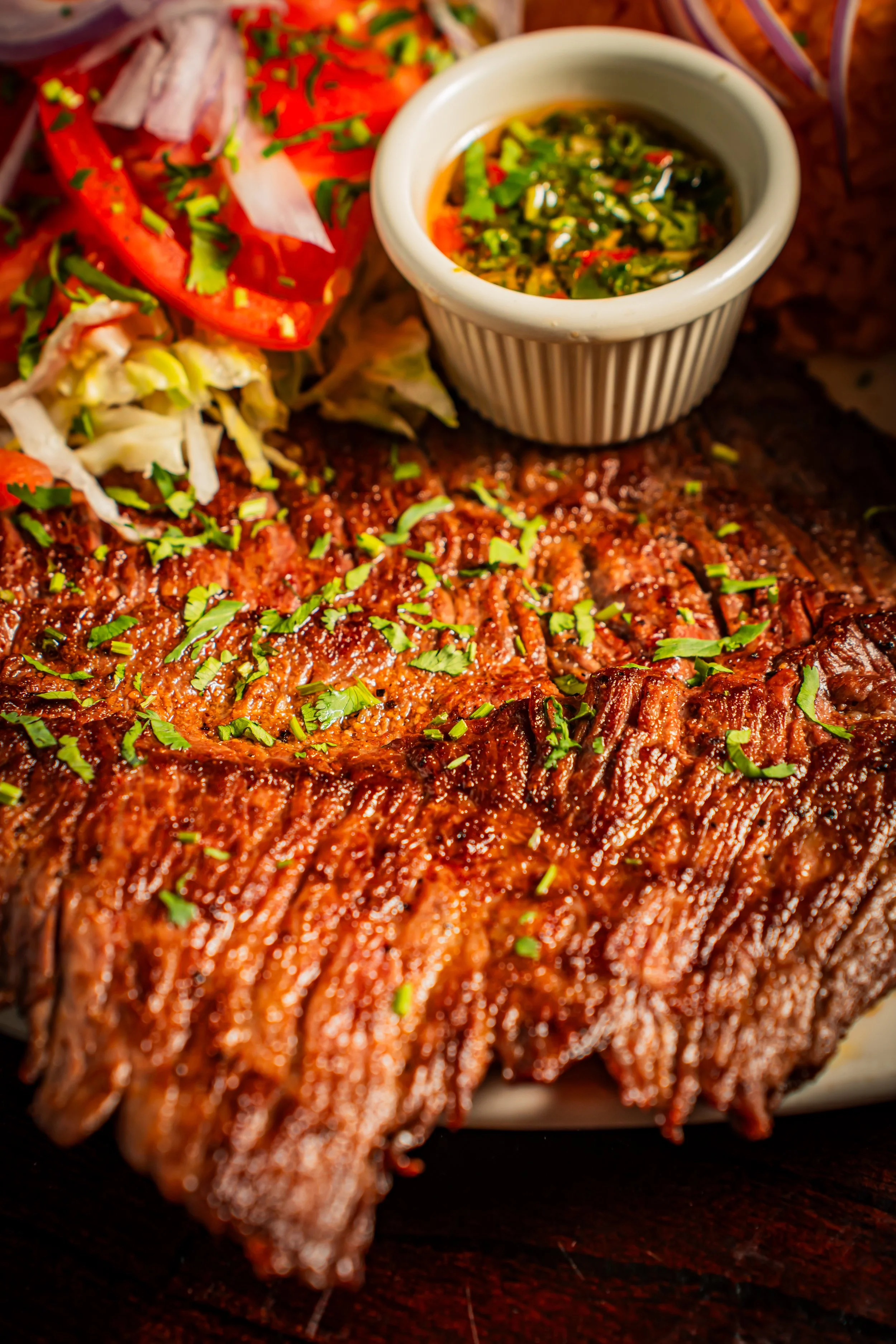Close-up of a barbecue plate featuring a large, glazed beef brisket garnished with chopped herbs, accompanied by sliced tomatoes, shredded lettuce, and a small ramekin of green sauce.
