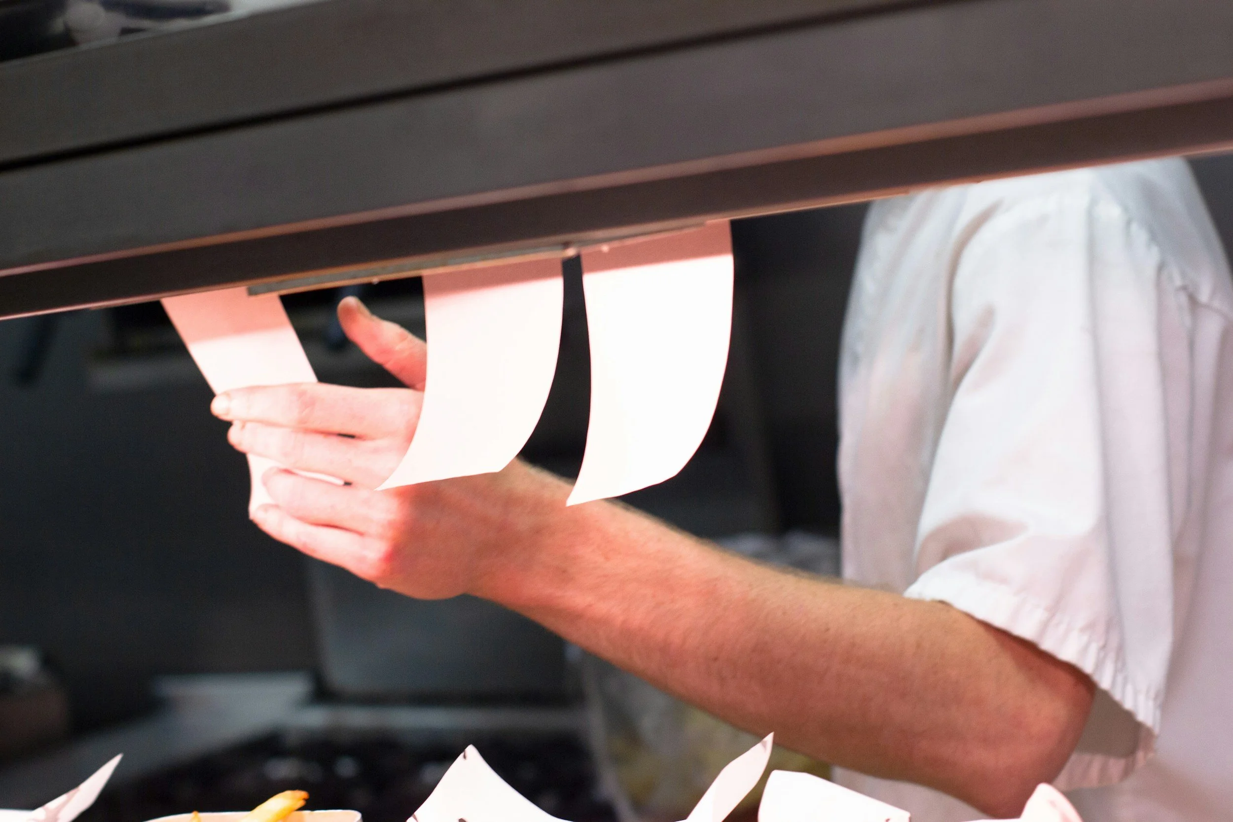 Close-up of a person making paper cones by hand, with a person's arm and partial torso visible, wearing a white shirt. The workspace appears to be a kitchen or paper preparation area with scattered paper pieces.
