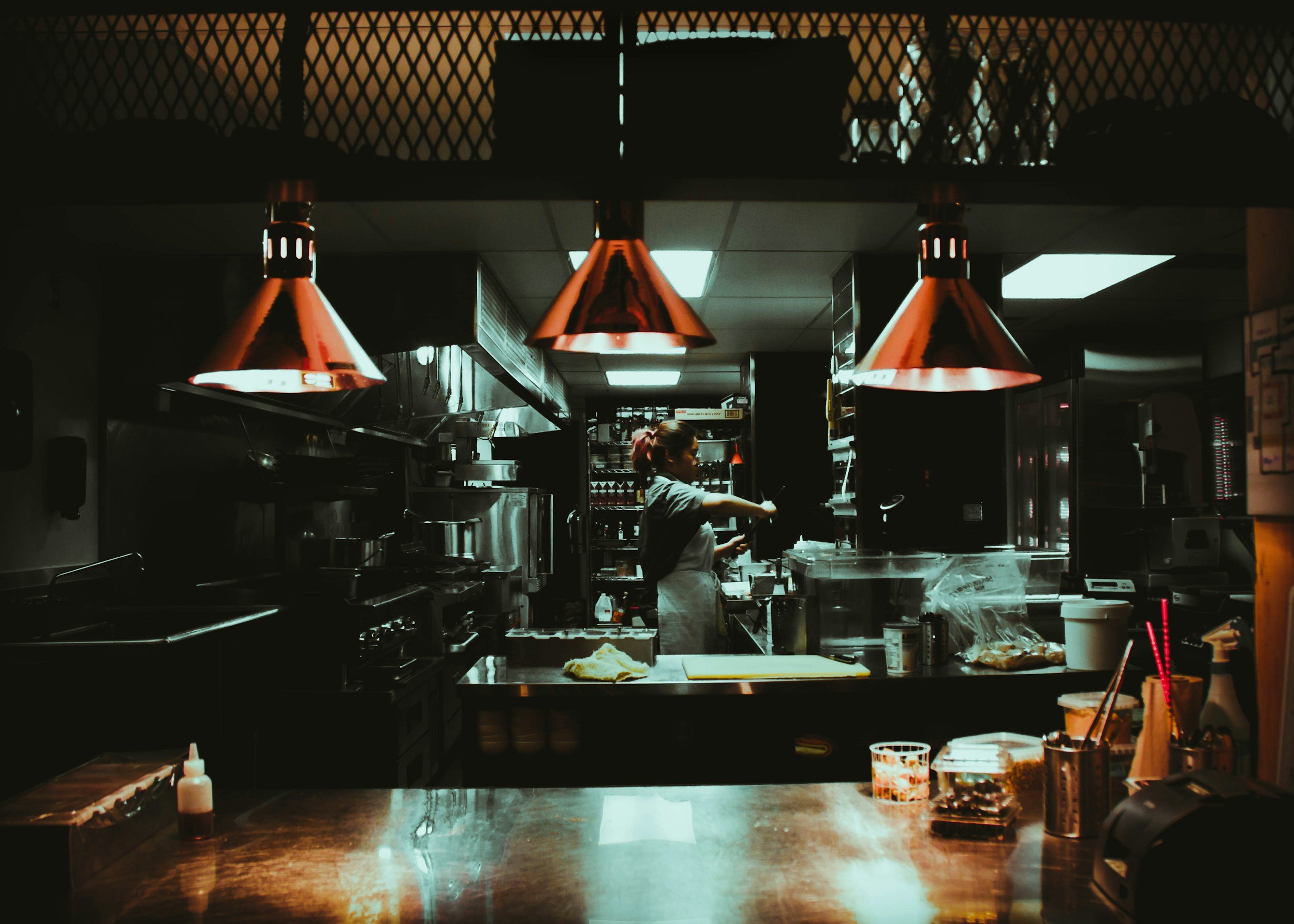 Inside a dimly lit restaurant kitchen, a female chef is preparing food, surrounded by kitchen appliances and utensils, with three red hanging lights above and a wooden counter in the foreground.