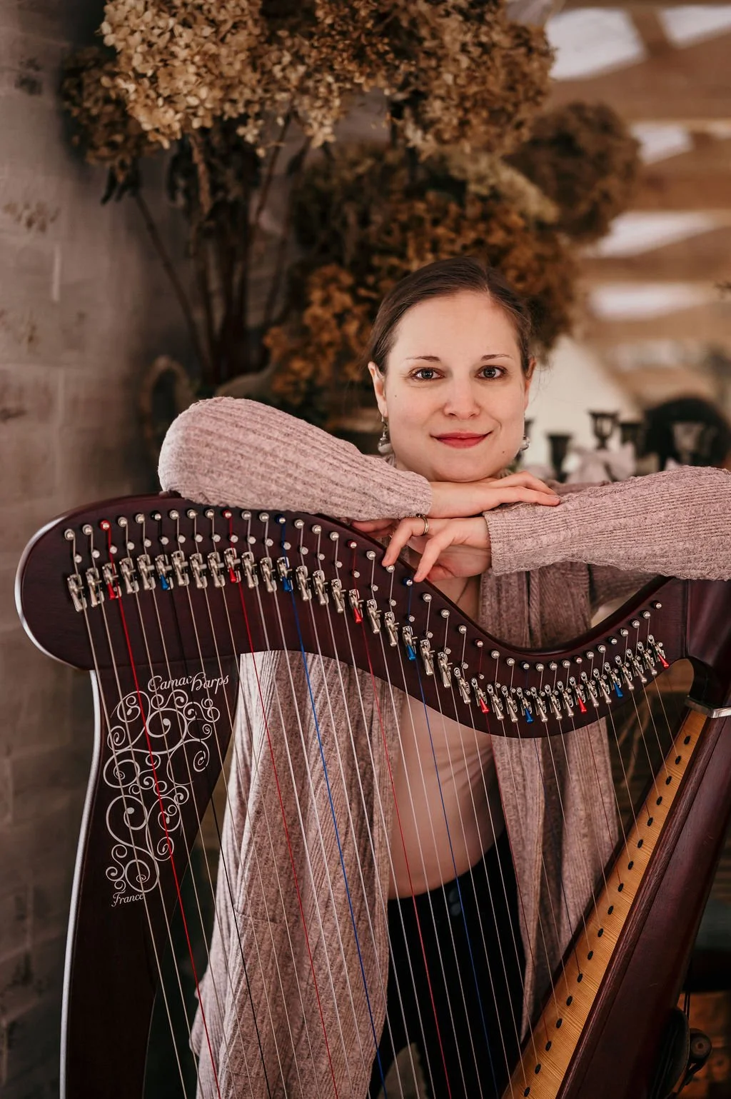 Une harpiste souriante pose en s'appuyant sur sa harpe, sur un fond décoré de fleurs séchées.