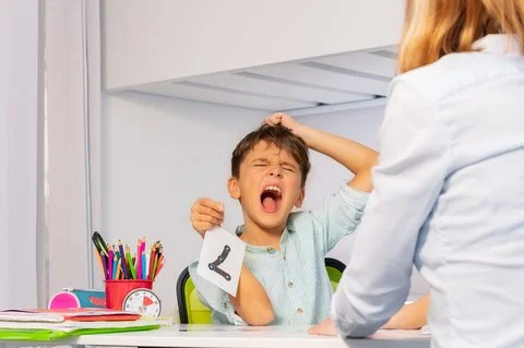 Une enfant ayant l'air frustrée ou en colère, assise à une table de dessin ou d'étude, dans un environnement intérieur, avec une adulte à côté.