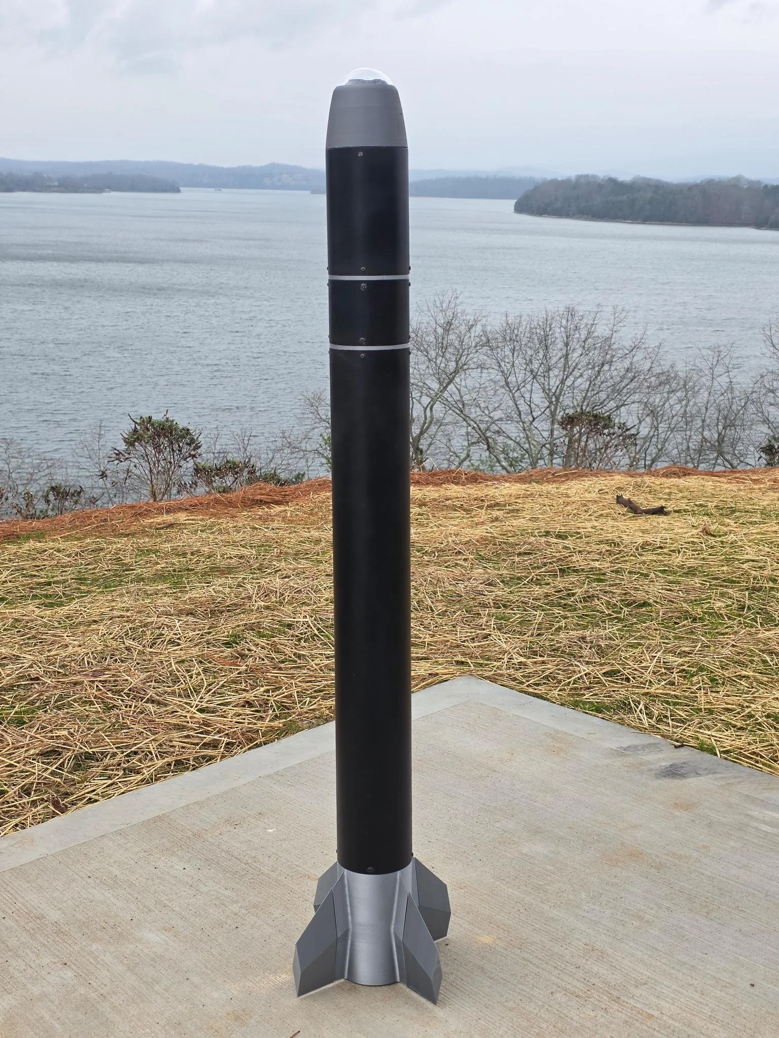 Model rocket standing on concrete platform overlooking a body of water with islands in the distance, trees, and overcast sky.