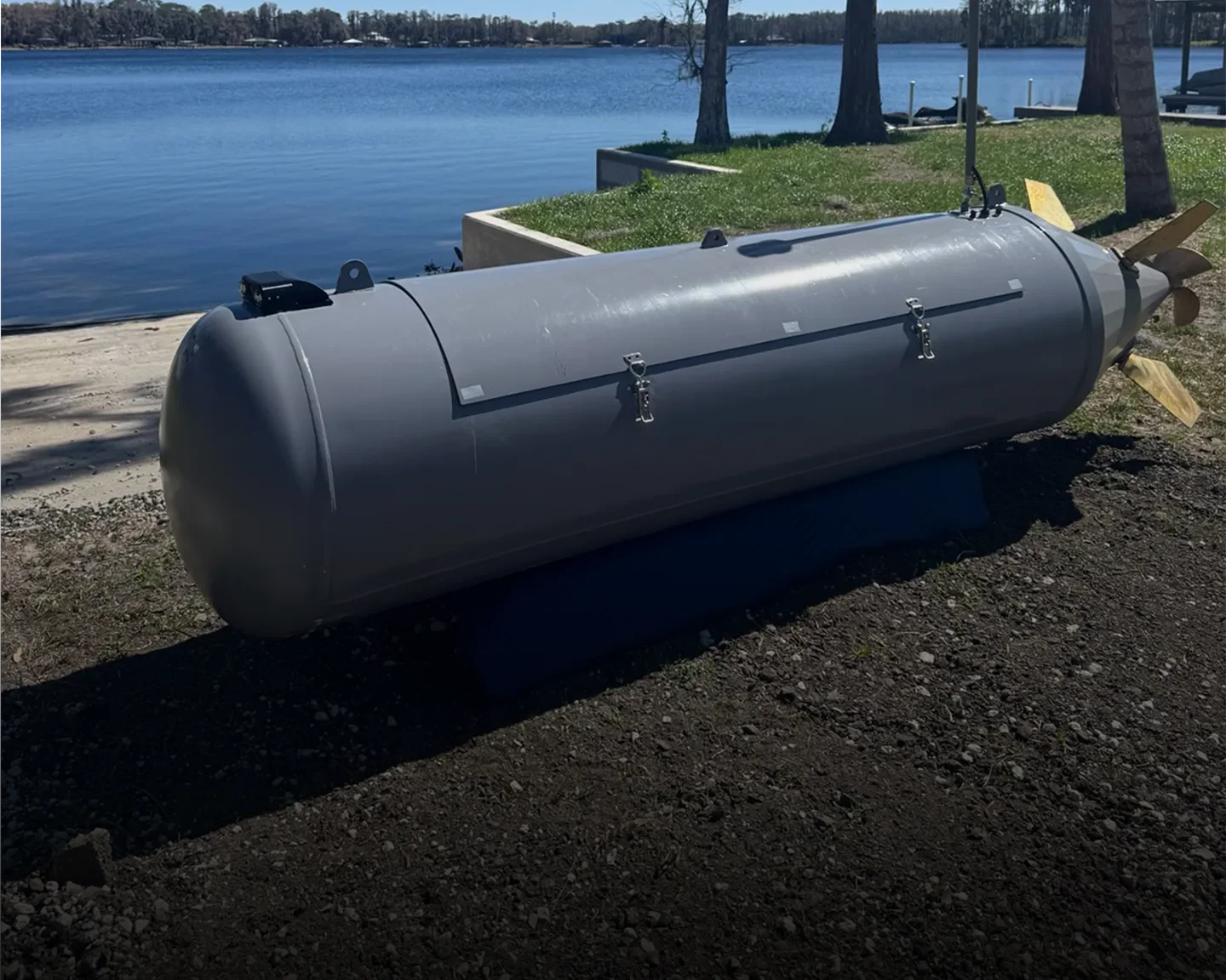 A large gray underwater device, possibly a torpedo or missile, rests on the ground near a lake, with trees and a dock visible in the background.