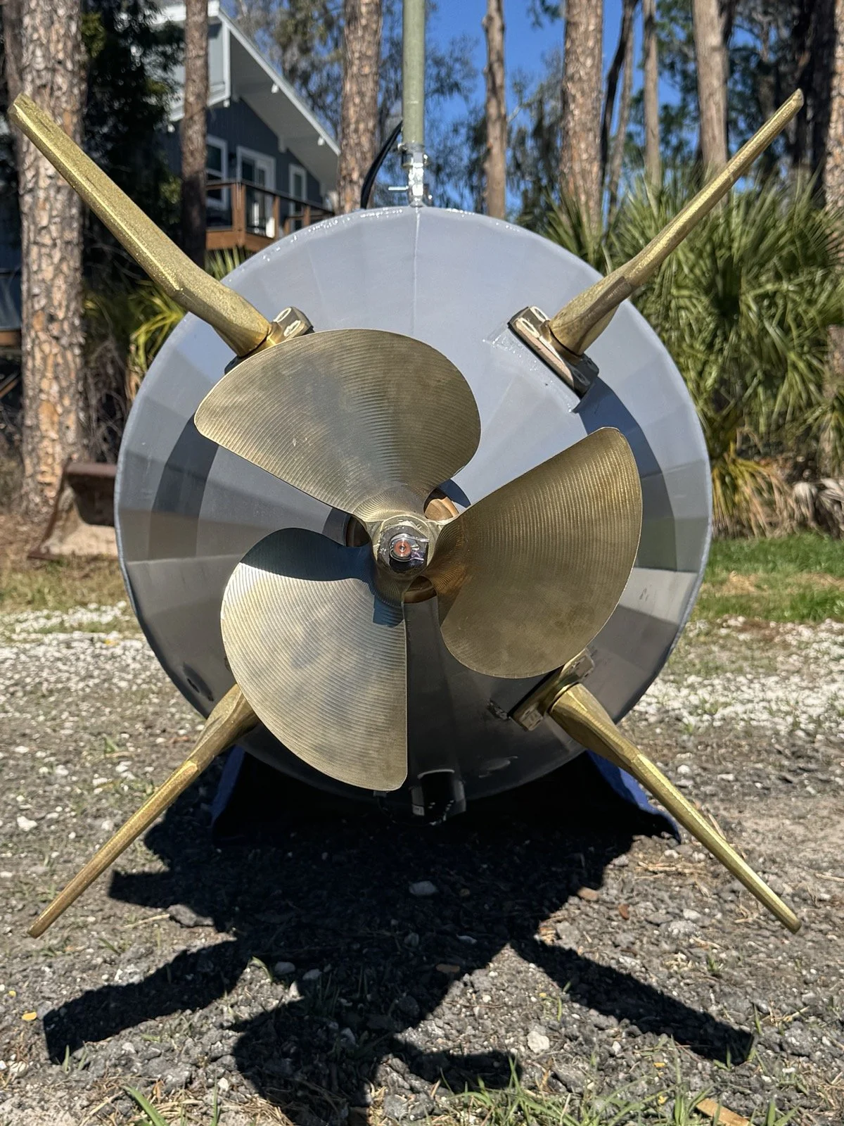 A model rocket with a sleek, gray metallic body and four golden fins on a grassy area with trees and a house in the background.
