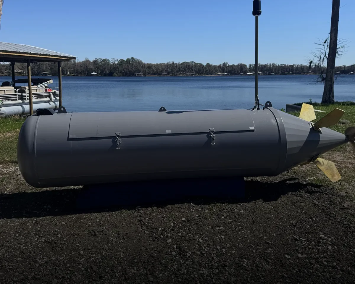 A large gray torpedo-shaped device with fins and a periscope-like antenna set on the ground near a lake, with trees and a dock visible in the background.