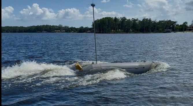 A small gray submarine on a body of water, with a thin antenna on top and trees in the background, under a partly cloudy sky.