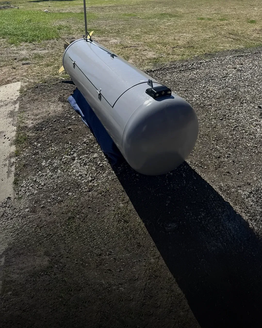 A cylindrical submersible resting on the ground outdoors, with a small black device on top and an antenna attached to one end, casting a long shadow on gravel and dirt.