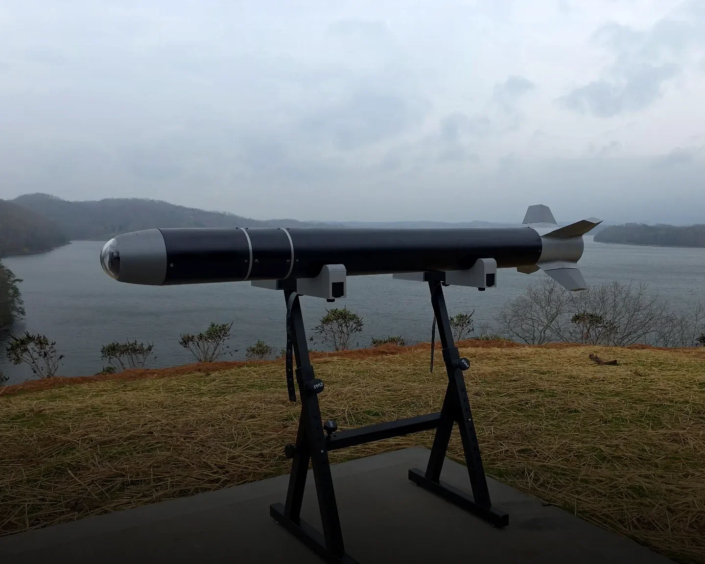 A model of a missile mounted on a stand overlooking a body of water with hills in the background under an overcast sky.