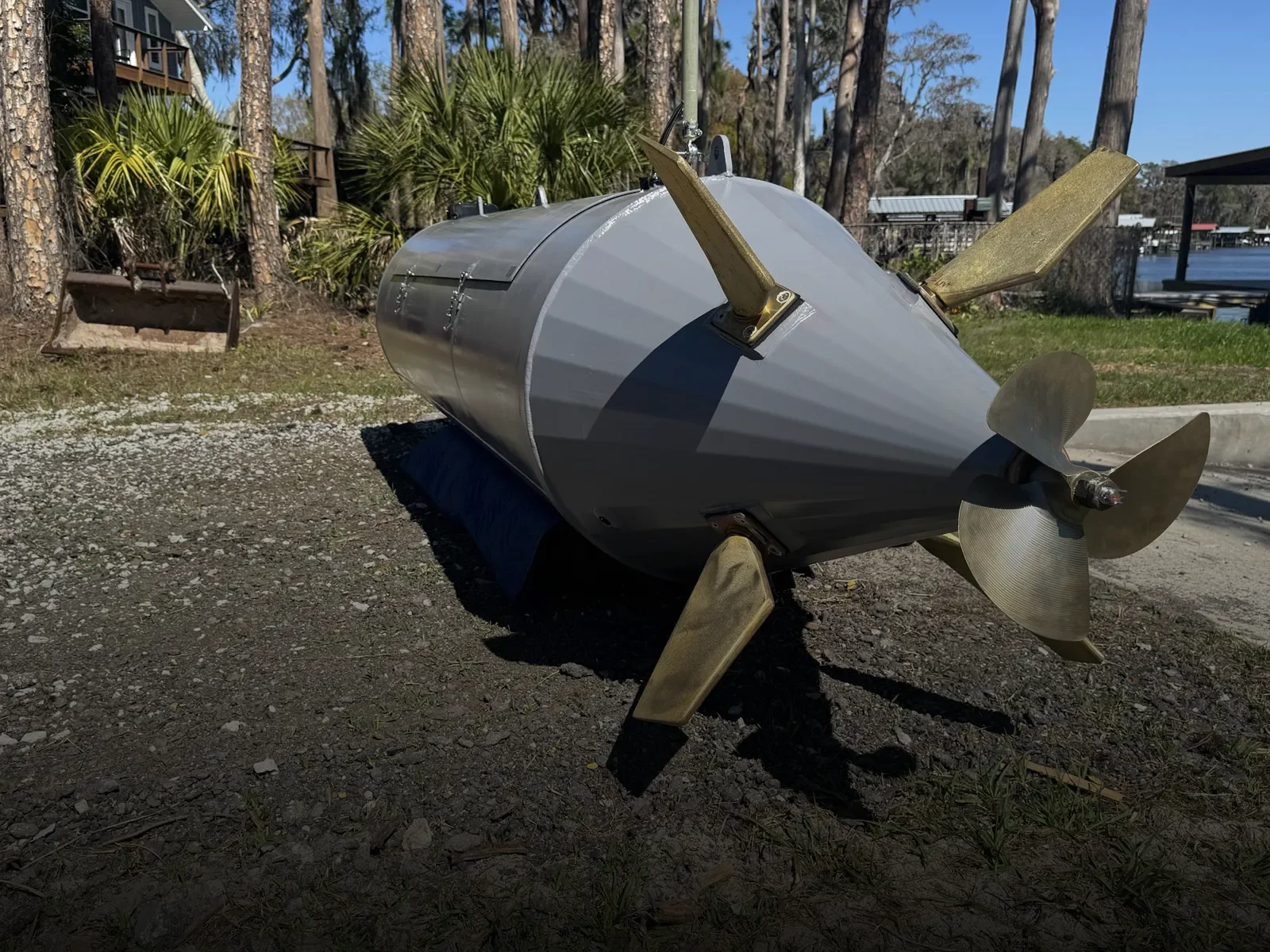 A model spacecraft with a cylindrical gray body, four angled gold-colored fins, and a three-bladed gold-colored propeller on the front, resting outdoors on a dirt surface with trees and a lake in the background.