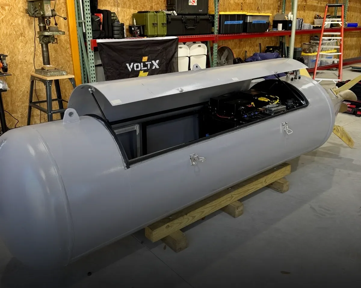 A large gray metal cylinder with a hinged top is open, revealing machinery inside, sitting on wooden supports in a workshop.