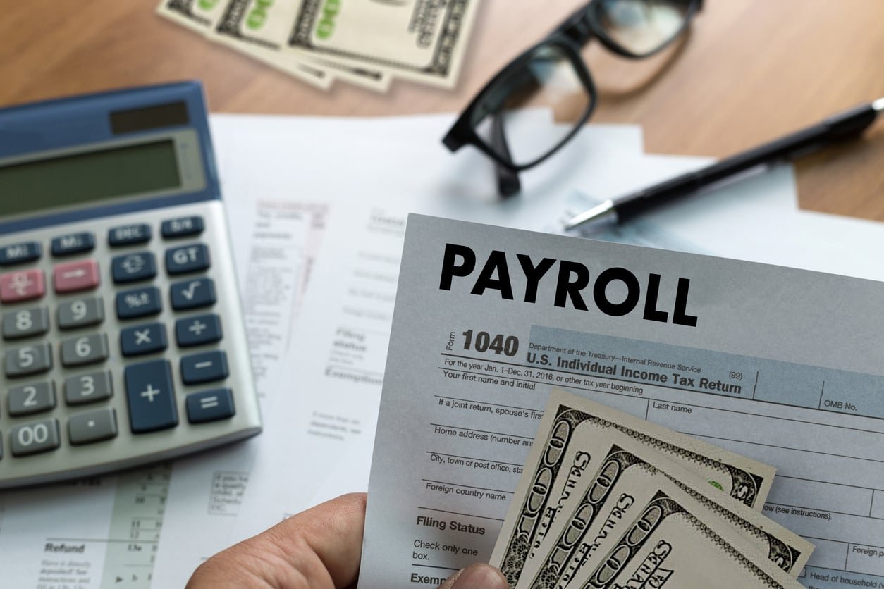A person holding a payroll form with cash and tax documents on a desk, along with a calculator, glasses, and a pen.