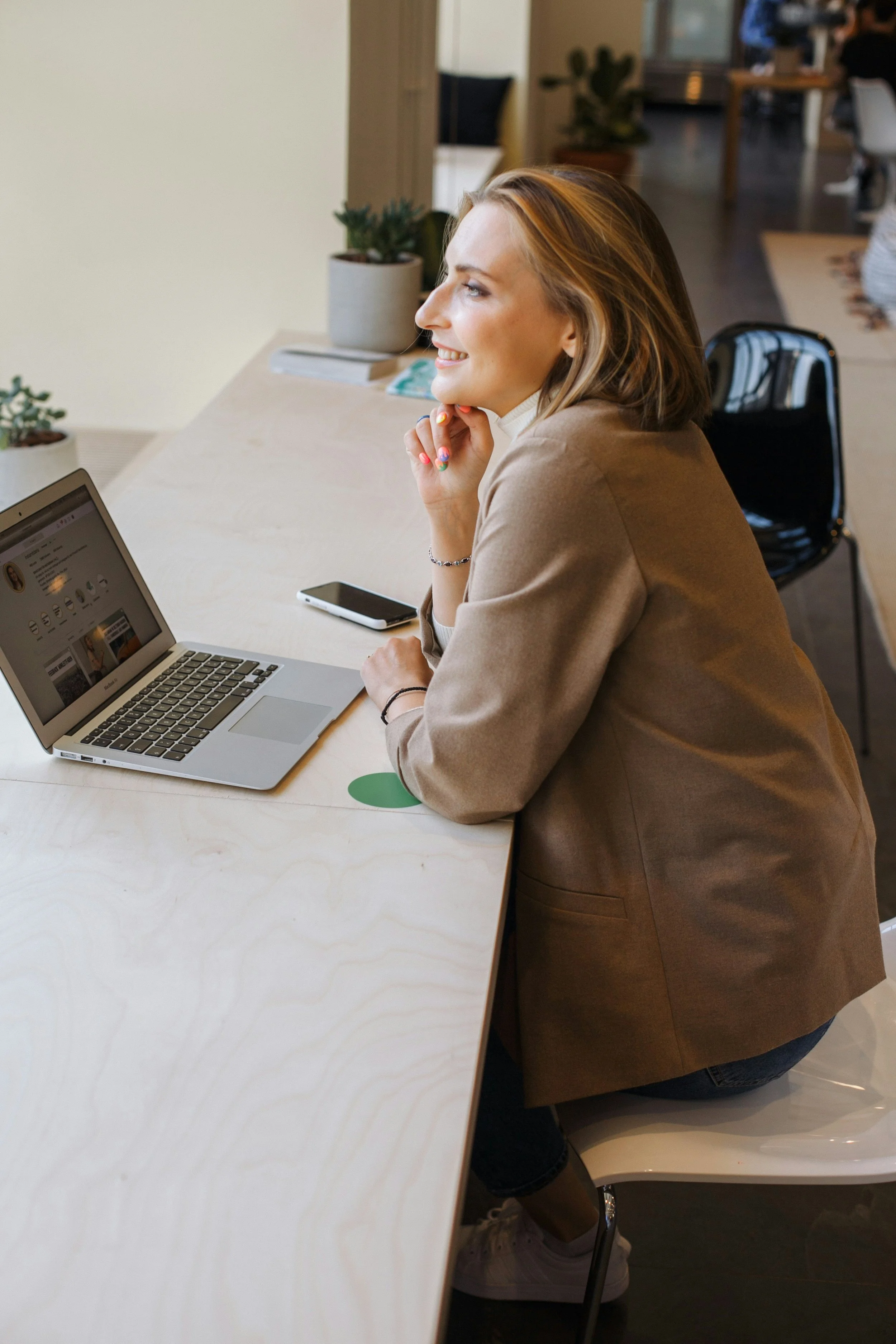 A woman sitting at a table in a cafe or workspace, smiling and looking to the side. She has shoulder-length hair, is wearing a beige blazer, and has colored nail polish. There is a laptop, a smartphone, and some potted plants on the table.