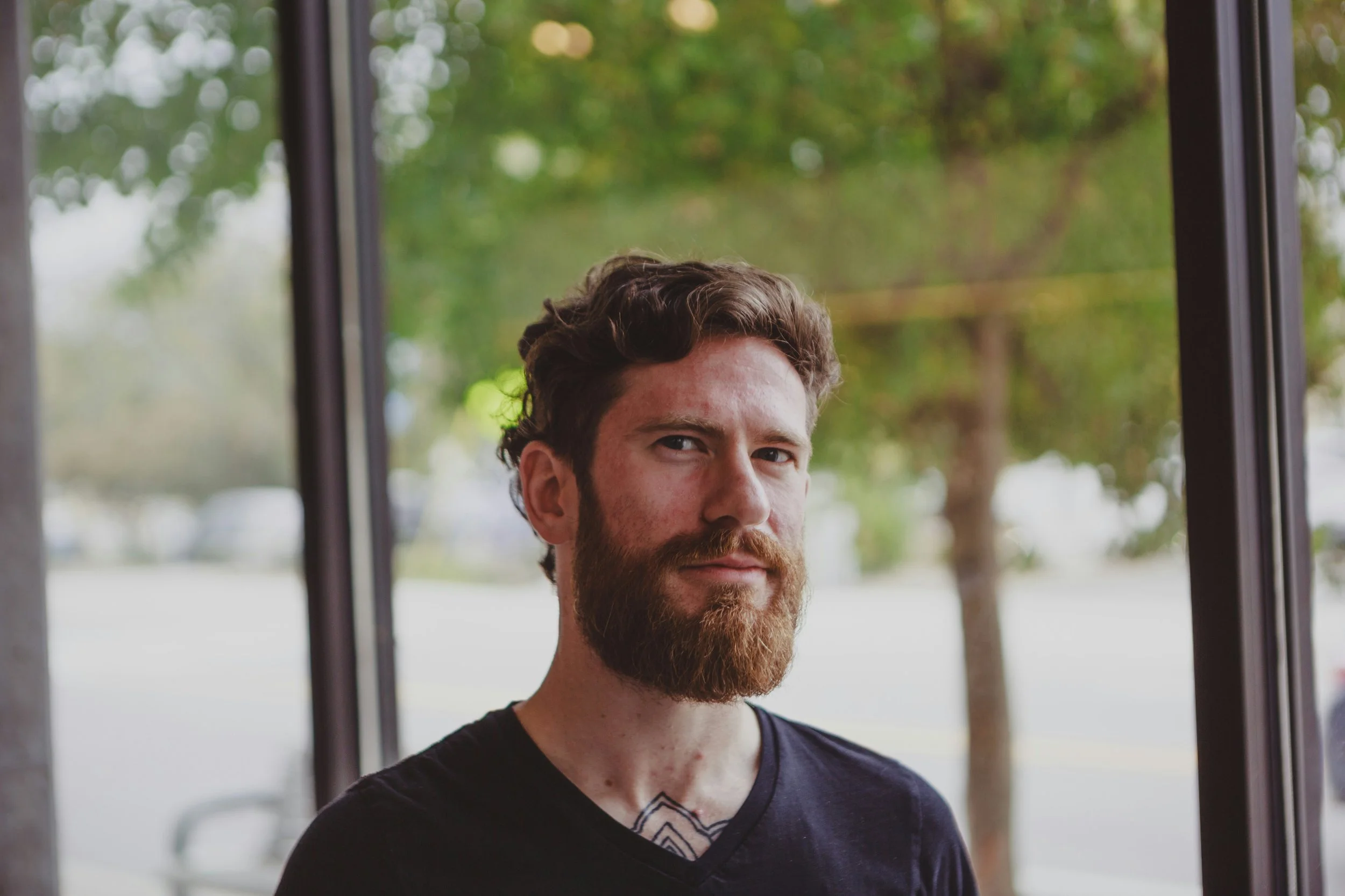 A young man with a full beard and curly brown hair is standing outdoors near a large window. He is wearing a black shirt with a V-neck and a patterned design visible near his collar. The background shows blurred trees and greenery.