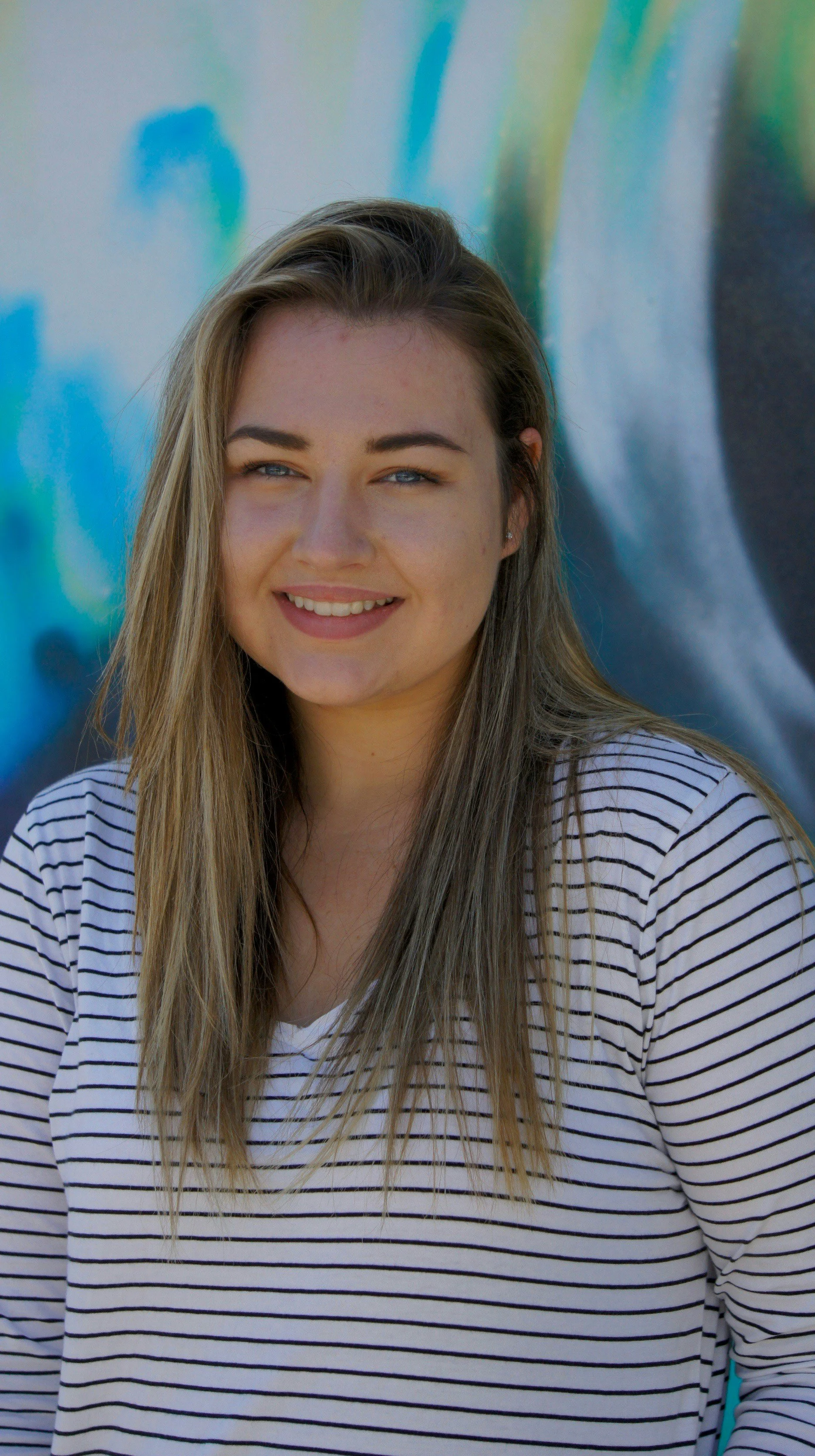 A young woman with long blonde hair and blue eyes smiling, wearing a white and black striped shirt, standing in front of a colorful graffiti wall.