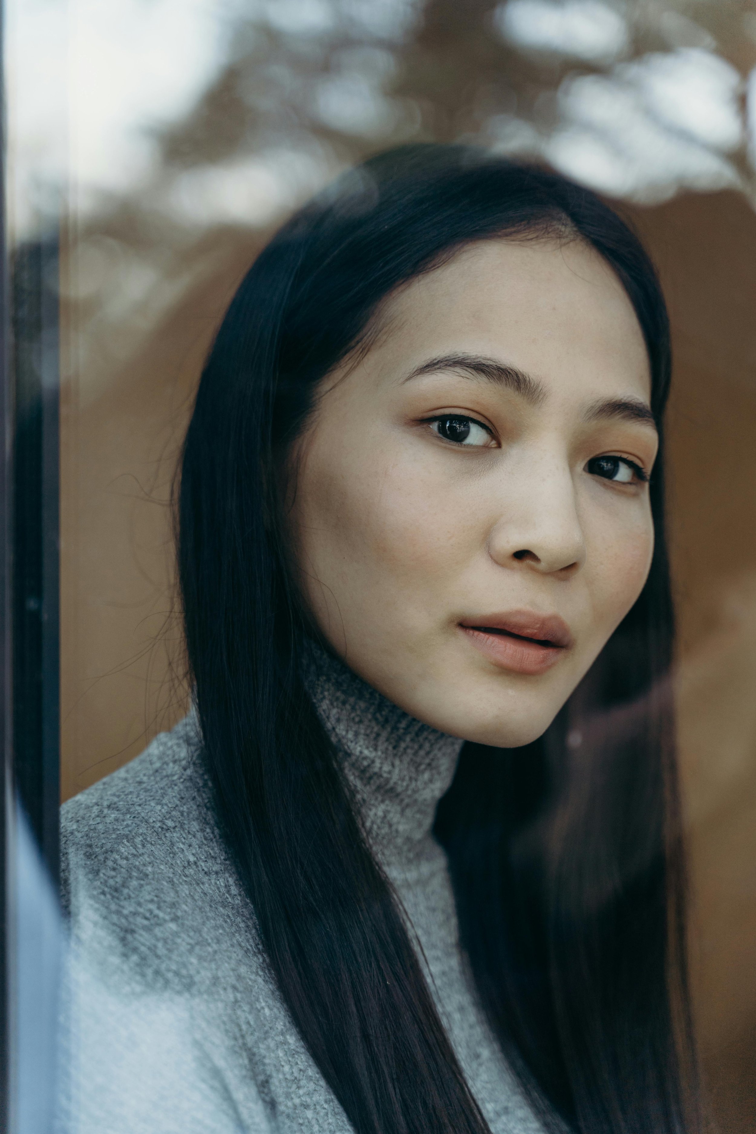 Close-up of a woman's face with long black hair, wearing a gray turtleneck sweater, looking at the camera with a calm expression.