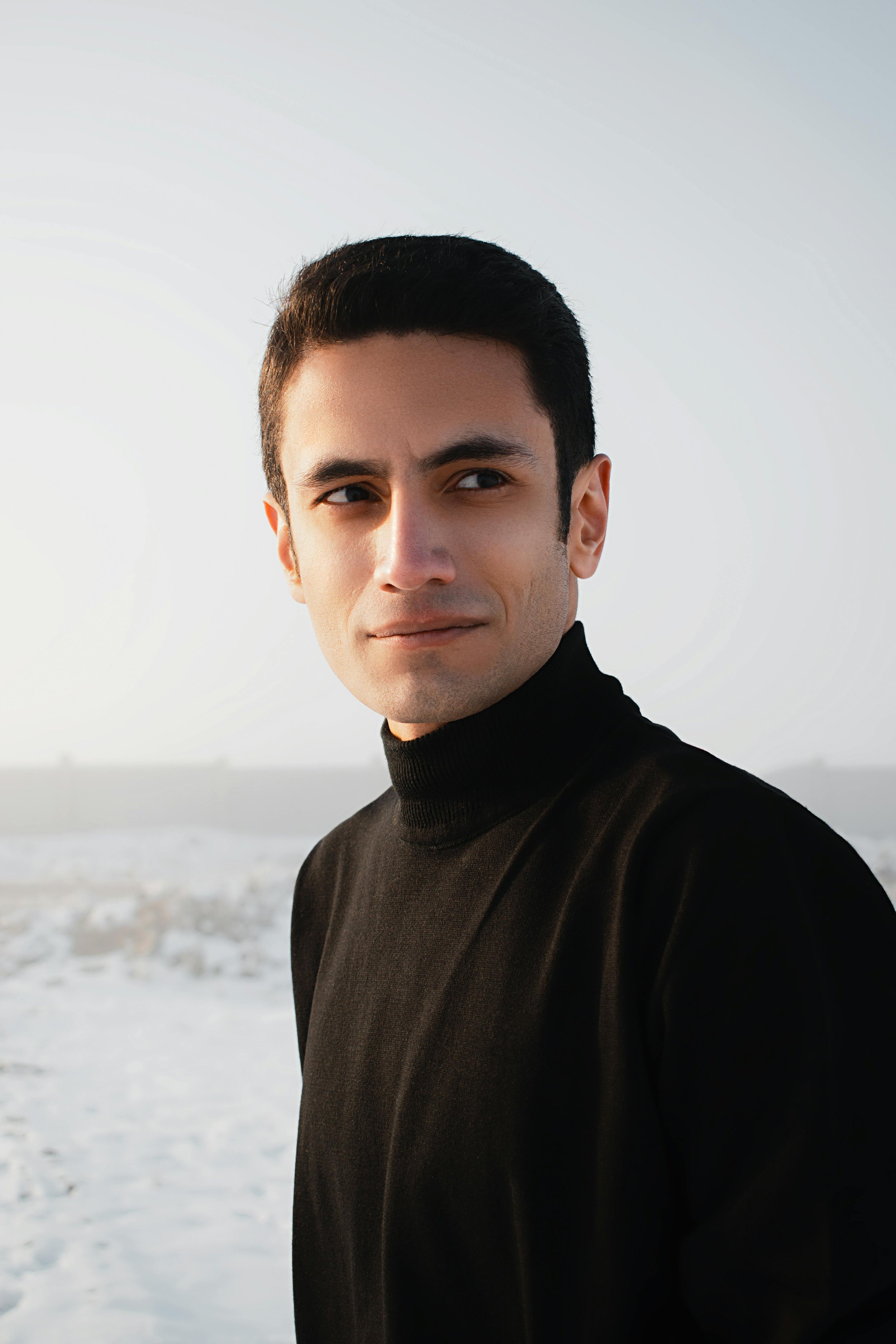 Young man with dark hair and light skin wearing a black turtleneck sweater, standing outdoors on a beach with complex cloud sky in the background.