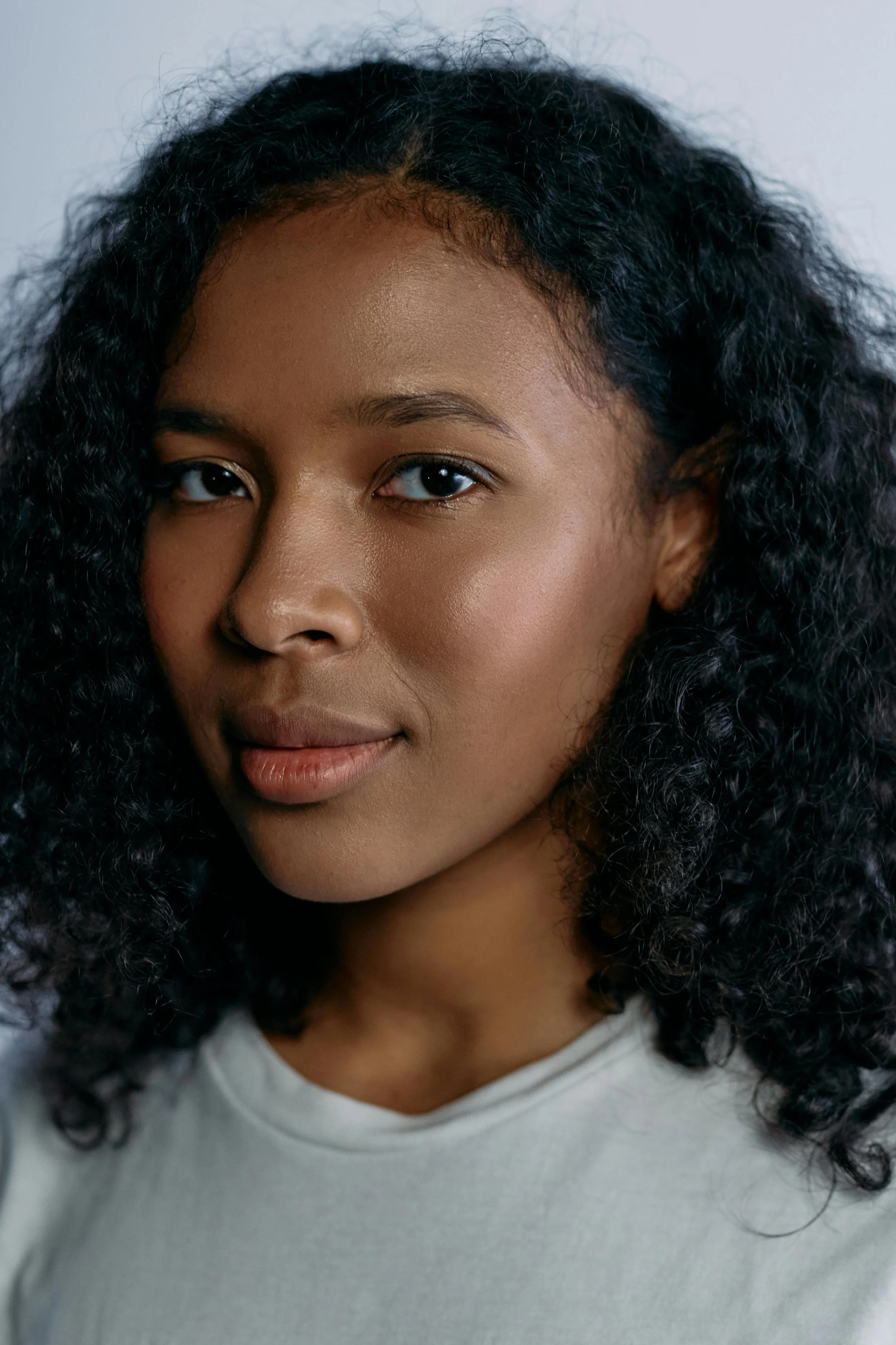 Close-up of a young woman's face with dark curly hair, smiling softly, wearing a light-colored shirt.