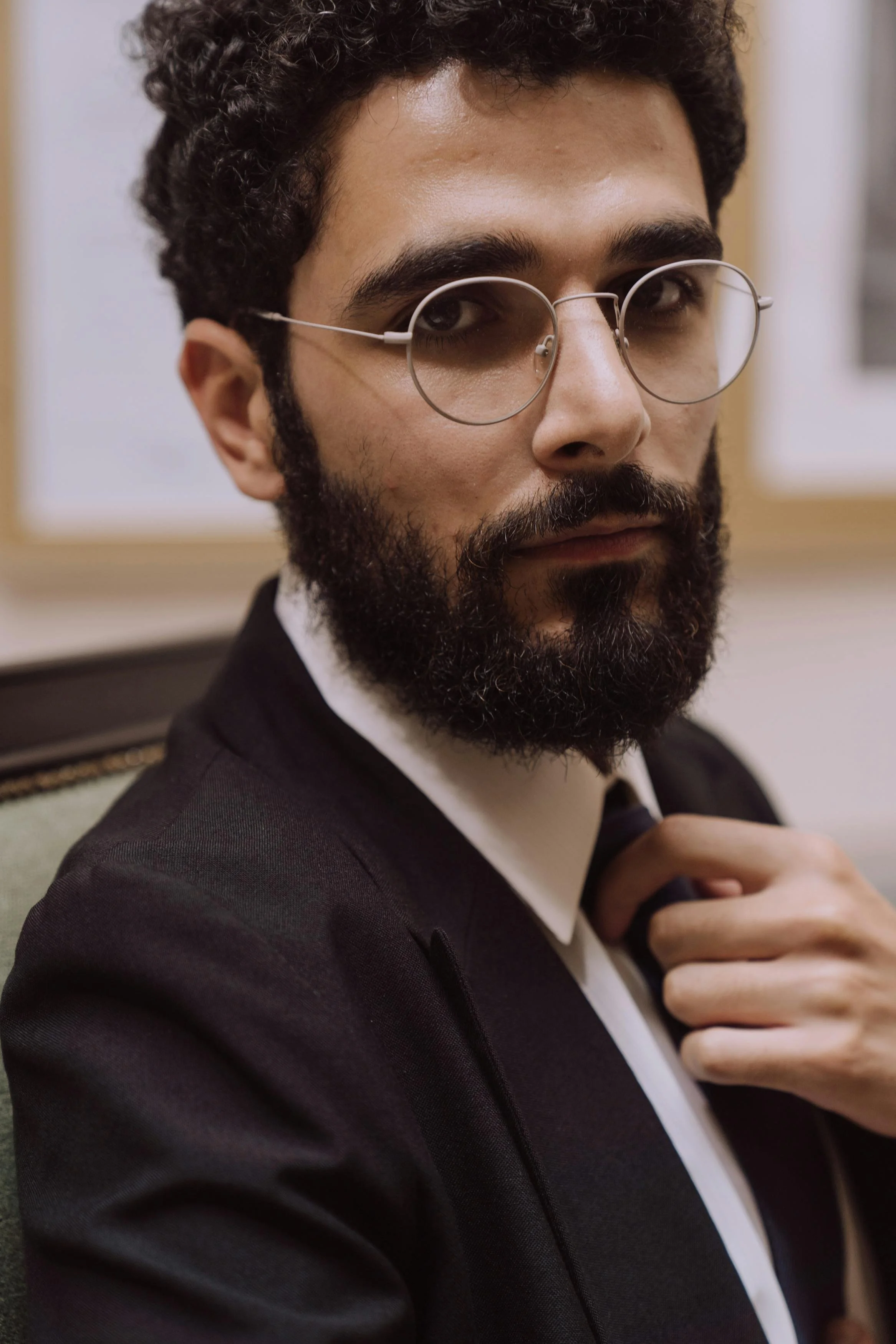 A close-up shot of a man with dark, curly hair and a beard, wearing round glasses in a black suit with a white shirt, sitting in front of a blurred background.