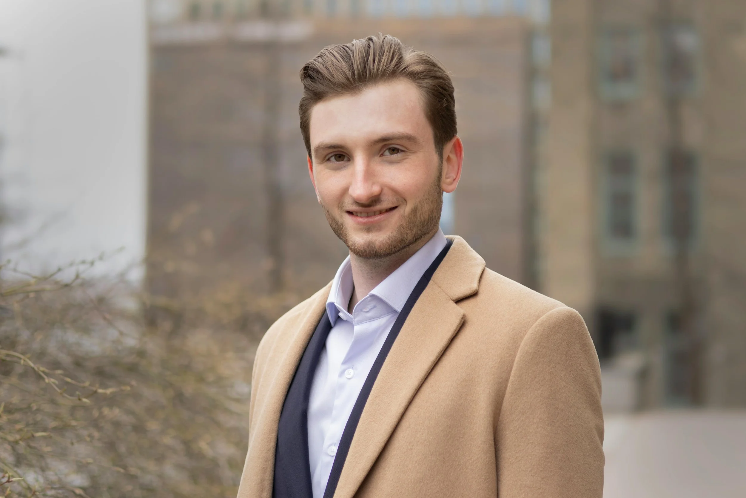 Young man outdoors wearing a beige coat, white shirt, and black tie, smiling with brown hair, on a cloudy day with buildings in the background.