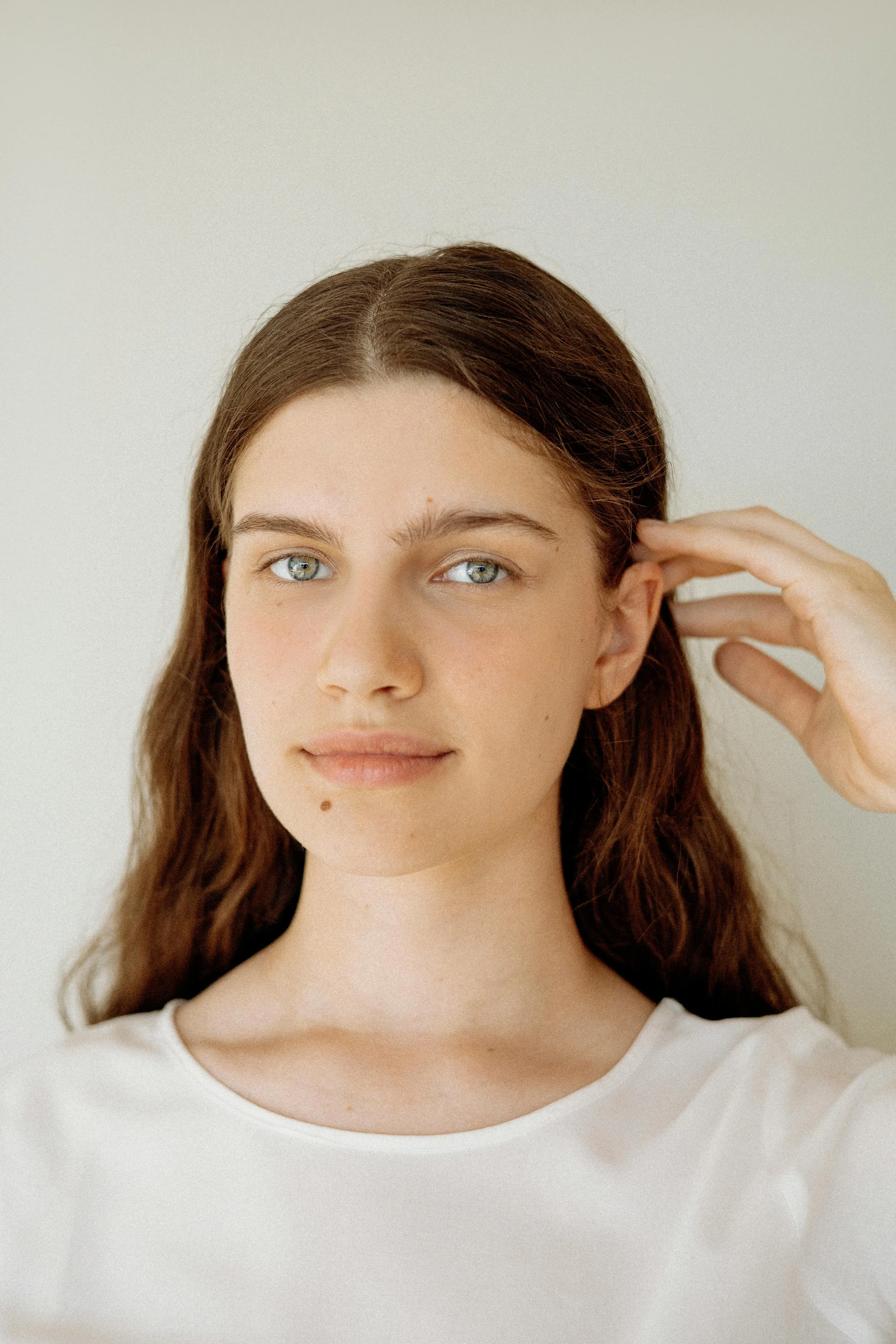 Close-up of a young woman with brown hair and green eyes, wearing a white top, touching her ear with her right hand against a plain, neutral background.