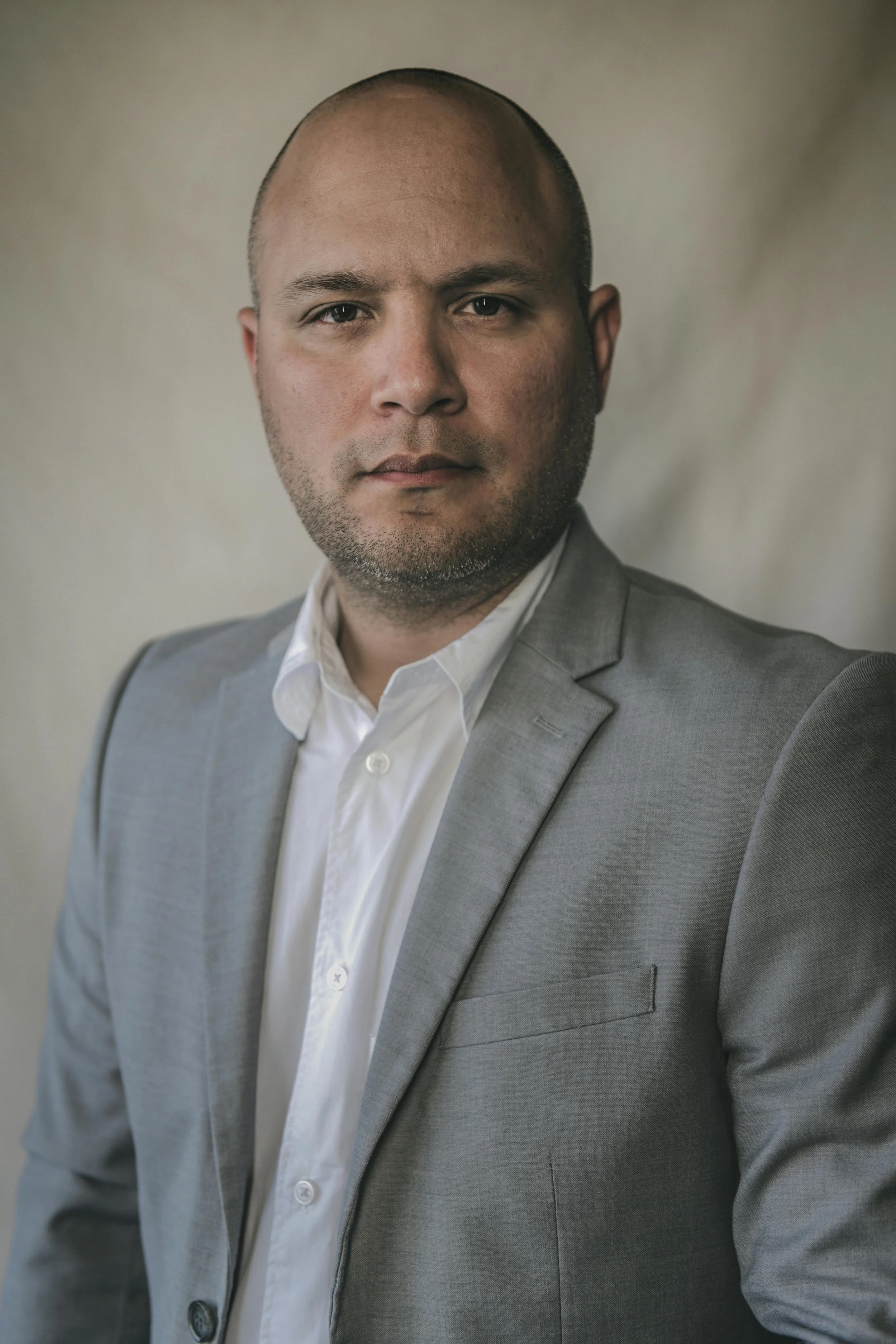 Portrait of a man wearing a light gray suit and white shirt, looking directly at the camera with a neutral expression, against a neutral background.