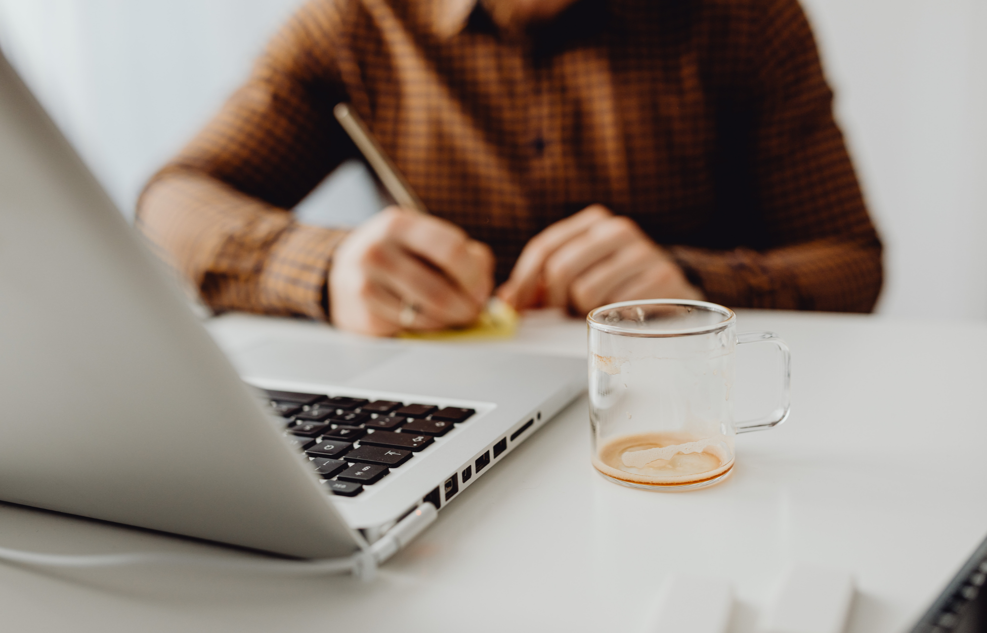 Person in brown checkered shirt writing at a desk with a laptop and a clear mug with coffee or tea