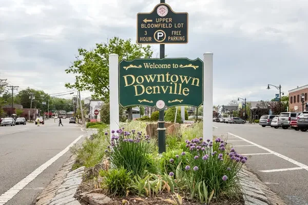 A green sign that says "Welcome to Downtown Denville" is in the center of a small landscaped area with purple flowers. Above it, there is a black parking sign indicating free parking for up to three hours at the Upper Bloomfield Lot. The background shows a street with parked cars, buildings, and cloudy skies.