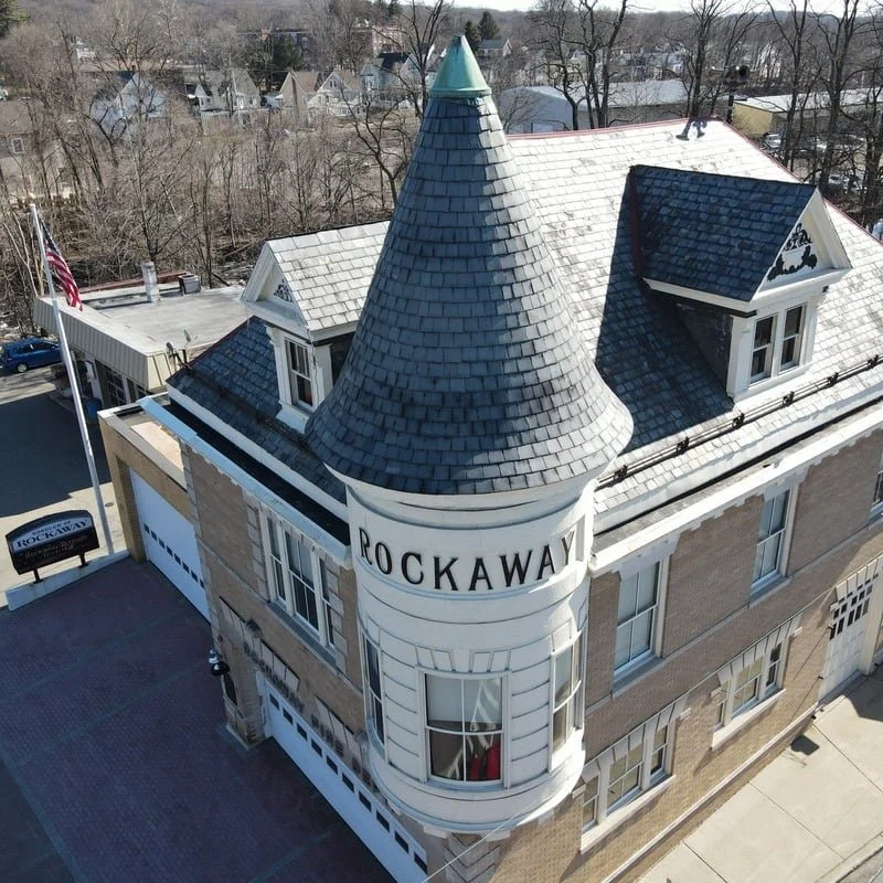 A three-story building with a turret and dormer windows, labeled 'ROCKAWAY', with a parking lot and American flag nearby, situated in a residential area with leafless trees.