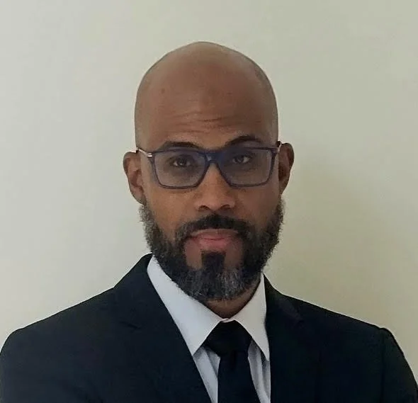 A man with a bald head, glasses, and a beard, wearing a dark suit and tie, standing against a plain light-colored wall.