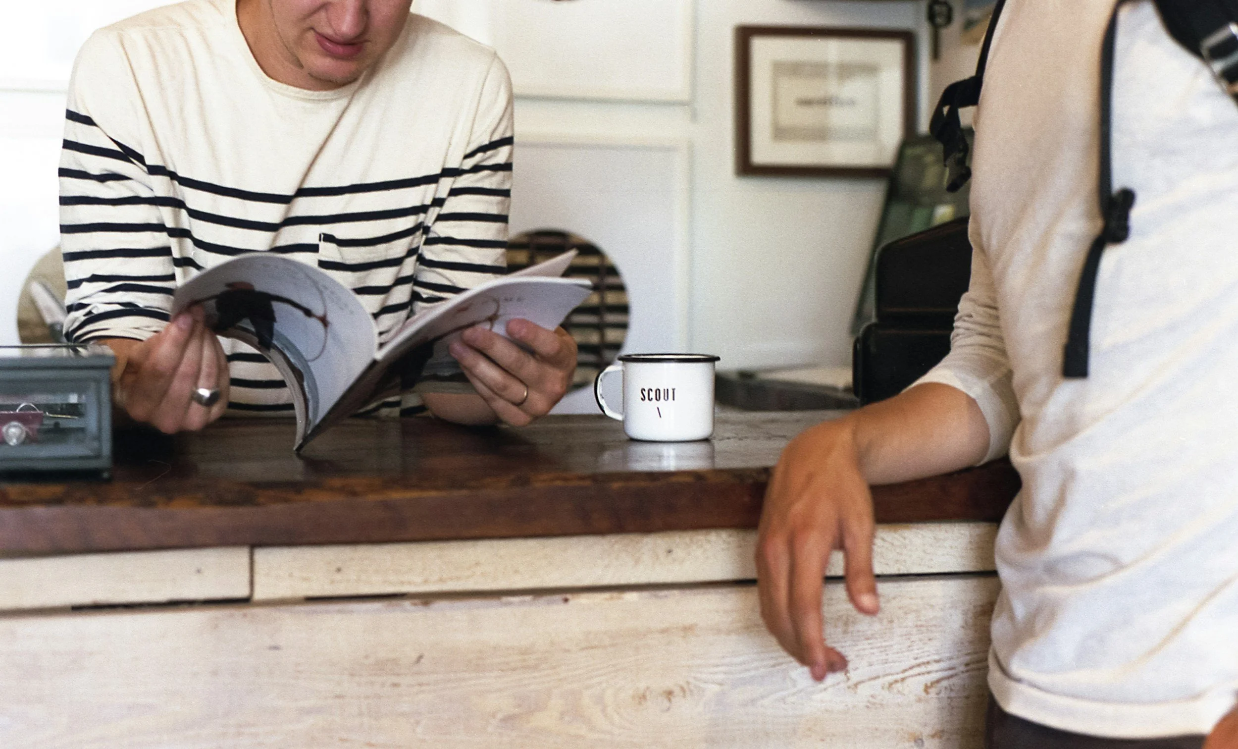 Pessoas sentadas na mesa, uma lendo uma revista, a outra com a mão apoiada sobre a mesa, com uma caneca branca com a palavra 'Scout' escrita. Ambiente interno com decoração simples, enquadramento focado nas mãos e parte superior dos corpos.