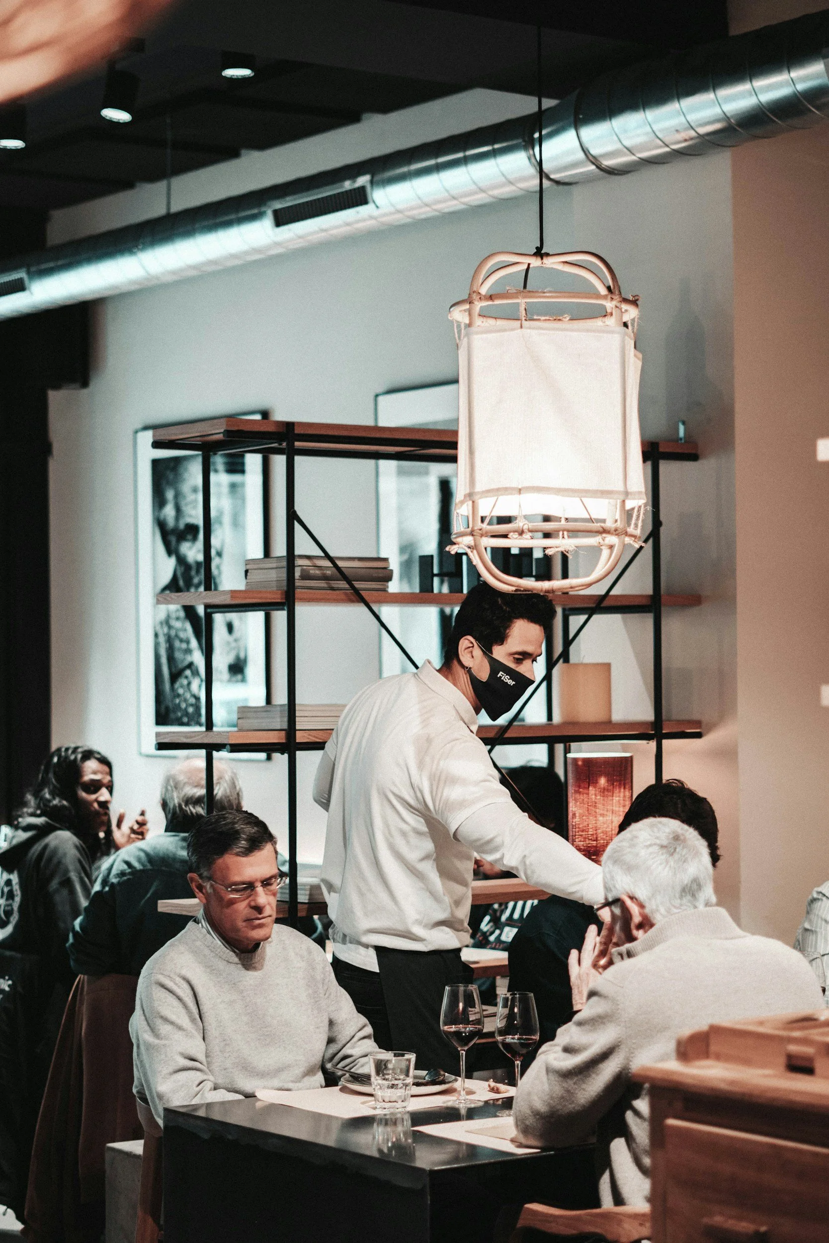 Cena em restaurante com várias pessoas. Homem com máscara atendendo clientes, alguns estão sentados em mesas com garrafas de vinho e copos. Decoração moderna com luminária e prateleiras.