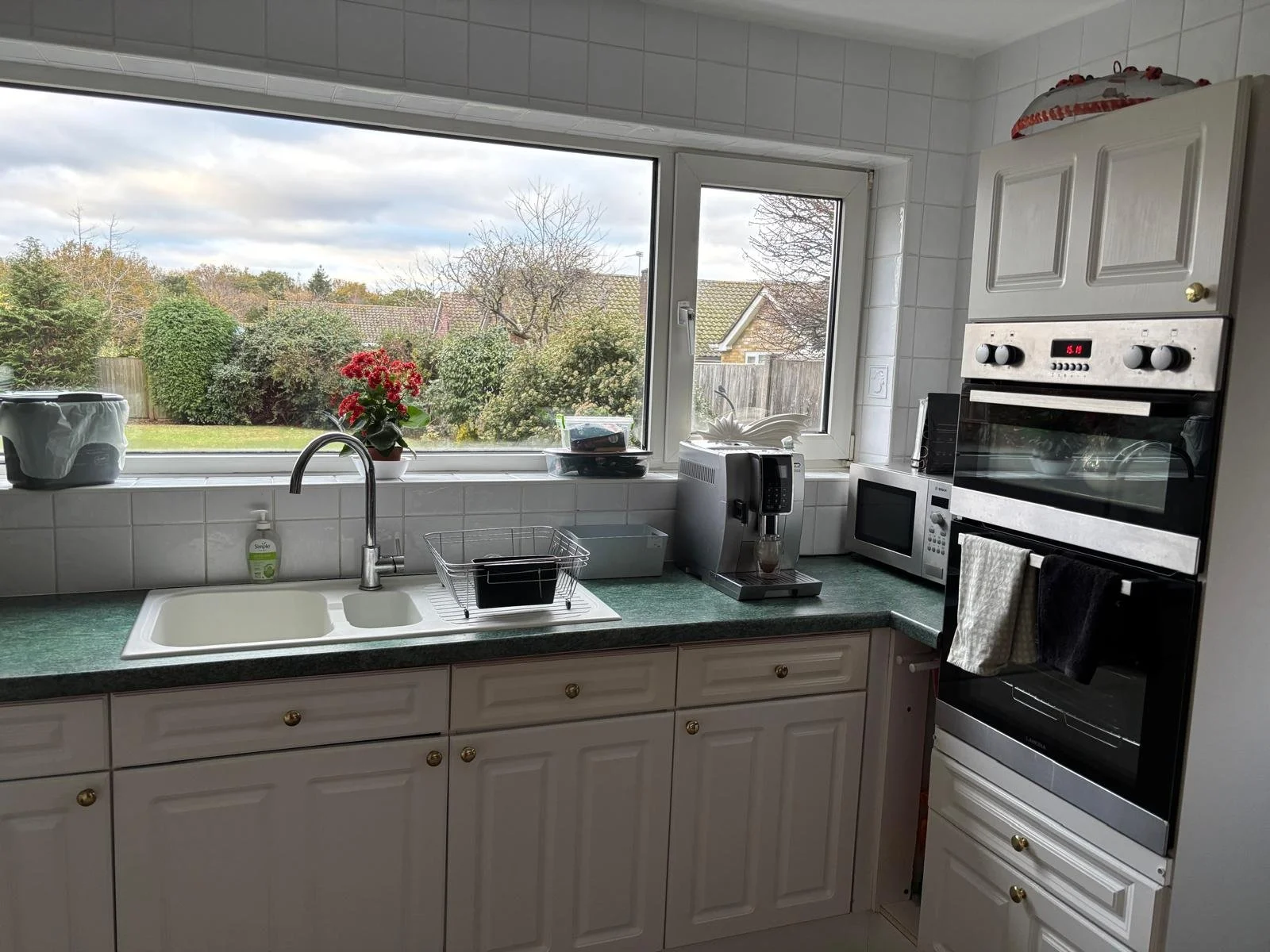 Kitchen with white cabinets, green countertop, coffee maker, microwave, oven, window with a garden view, potted plant, dish rack, soap dispenser.