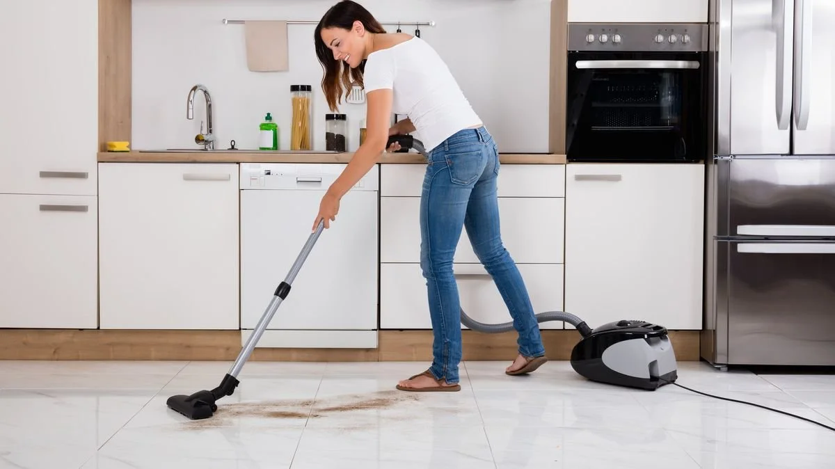 A woman vacuuming spilled dry food or dirt from a kitchen floor.