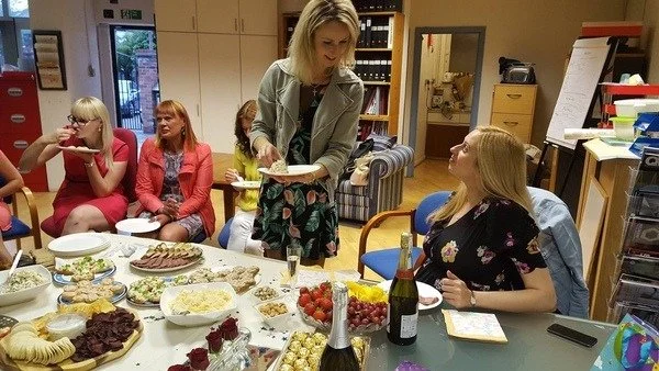 Four women gathered around a table with various foods, one woman serving food to others in an office setting.