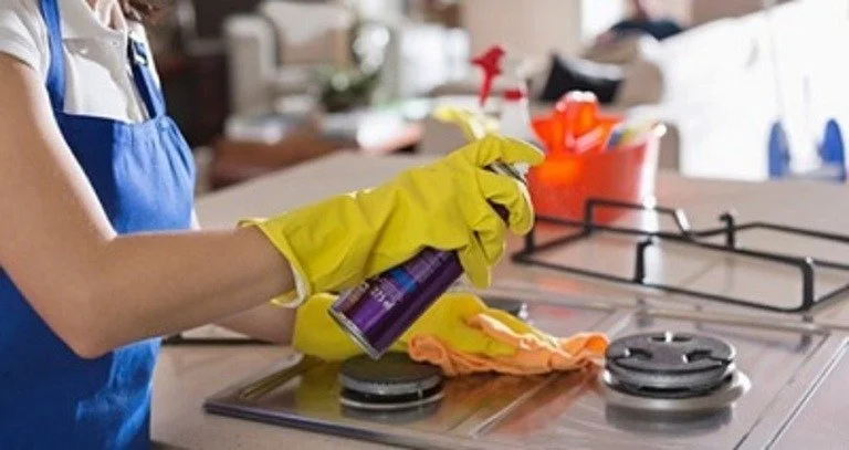 Close-up of a person wearing yellow rubber gloves cleaning a stove with a spray bottle and cloth.