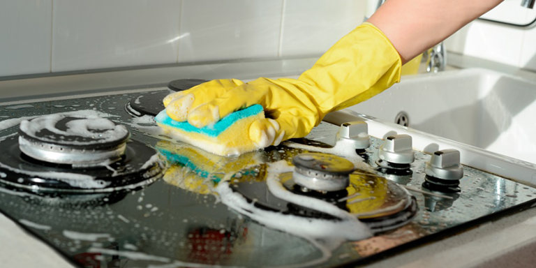 A person wearing yellow rubber gloves cleaning a stove with a sponge covered in soap suds.