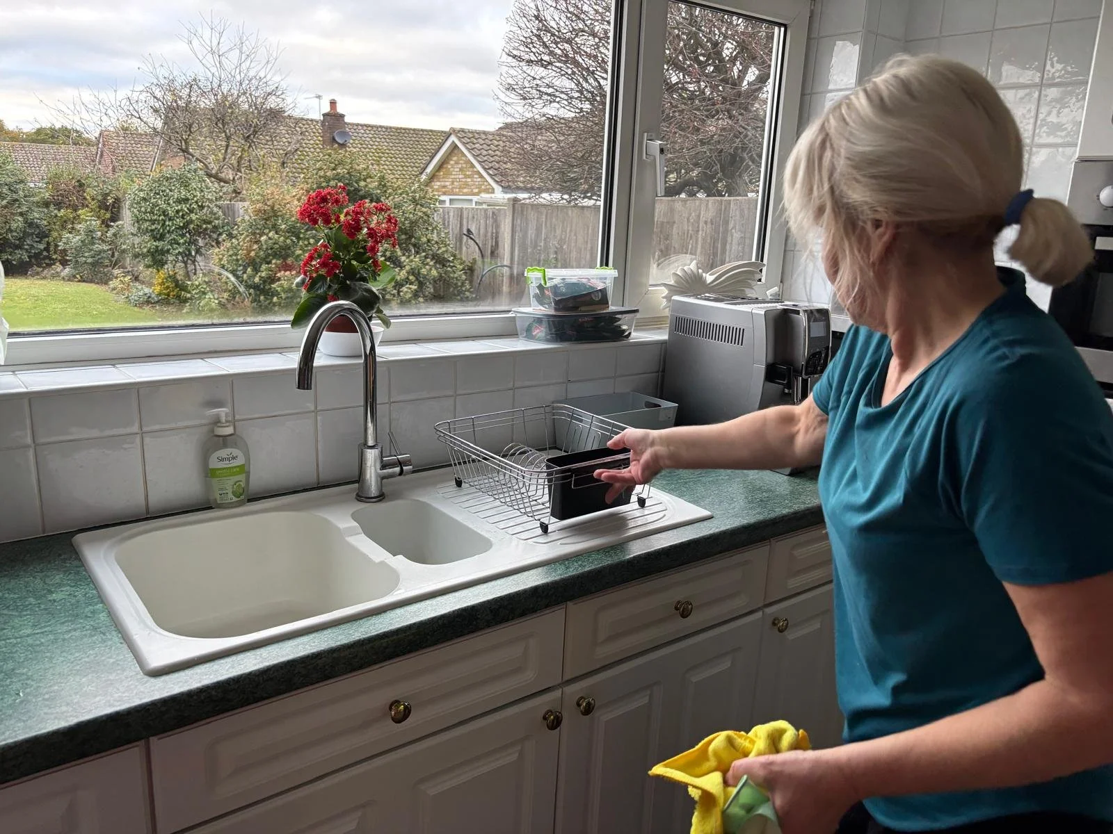 A woman with blonde hair tied in a ponytail, wearing a blue shirt, is drying dishes in a kitchen near a window with a view of a garden and neighboring houses. There is a potted red flower on the window sill, and kitchen appliances and dish racks are 
