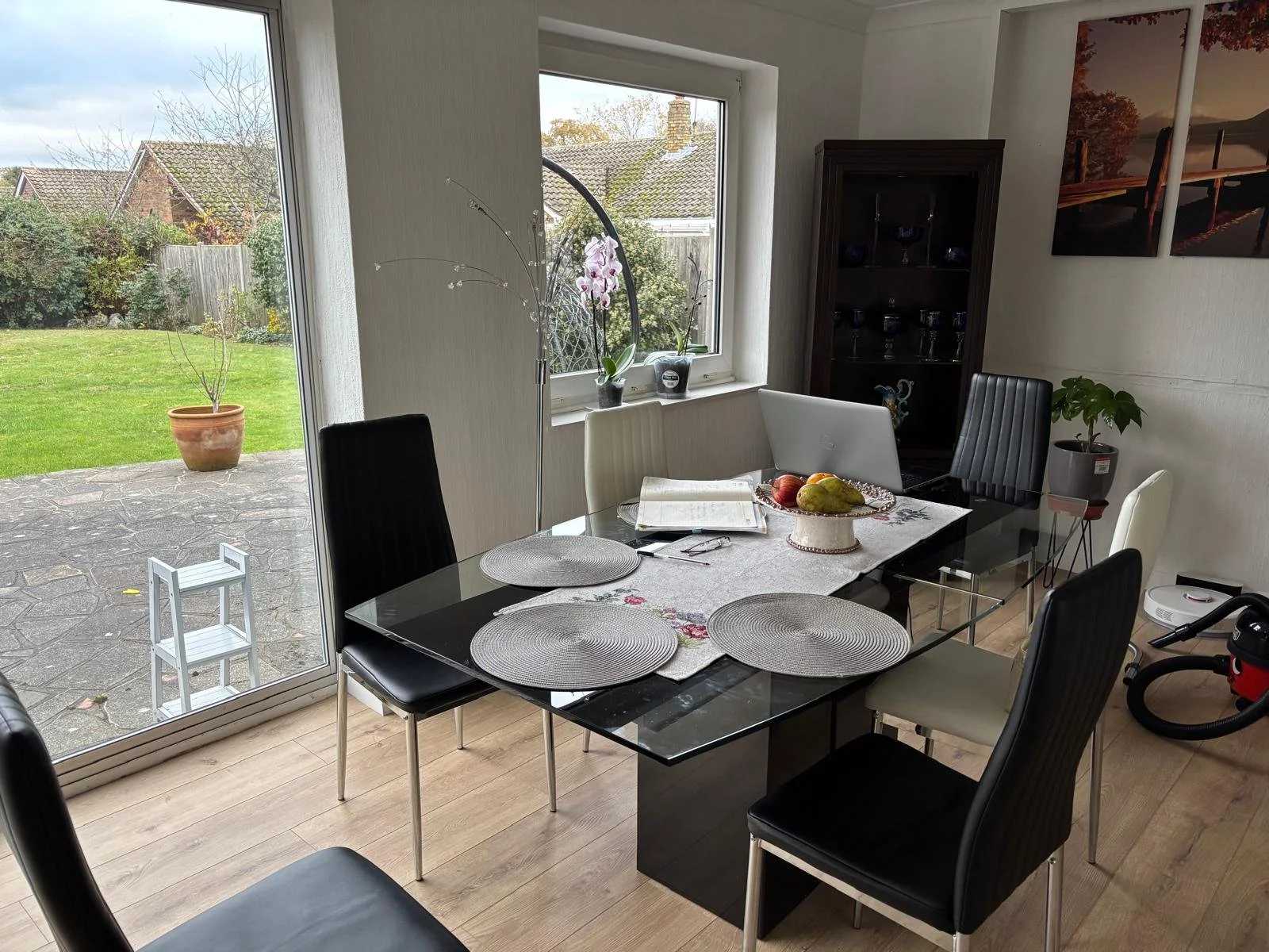 Dining room with a glass table set with placemats and a fruit bowl, surrounded by black and white chairs, with a large window showing a garden outside, and a cabinet with glasses, including decorative items and artwork on the wall.